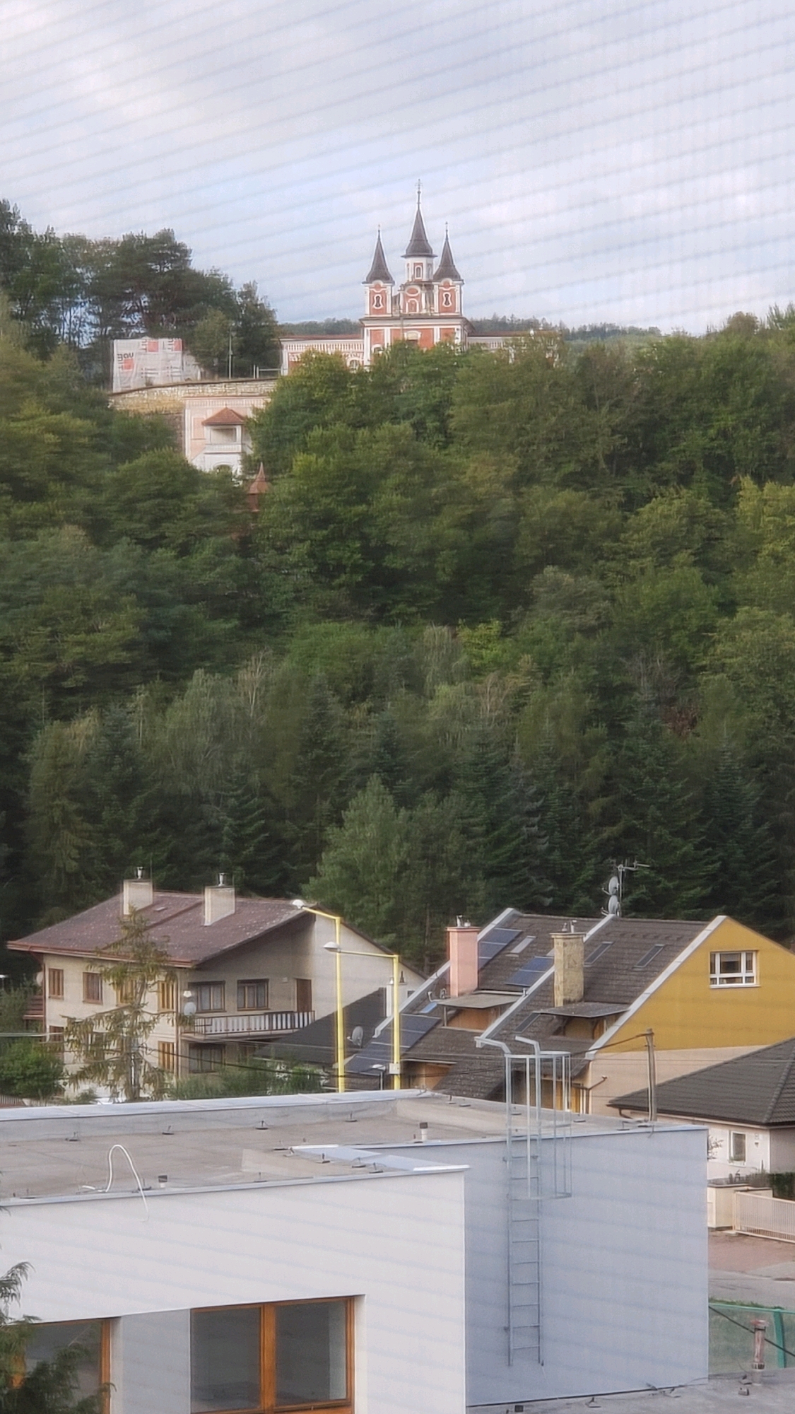 The image shows a view of a hilltop with a building featuring multiple spires, possibly a church or monastery, set against a cloudy sky. The building is red and white with dark spires. Below the hilltop, dense green trees cover the landscape. In the foreground, there are several residential buildings with tiled roofs and various architectural features, including windows, balconies, and chimneys. One building is yellow, and another is a lighter color. A white, modern structure with a flat roof and large windows is in the immediate foreground. A metal fire escape ladder is attached to the side of this building. The scene appears to be an urban or suburban area with a natural landscape backdrop. The location is identified as Prešov, Slovakia, likely from an elevated viewpoint. There are no people visible in the image.