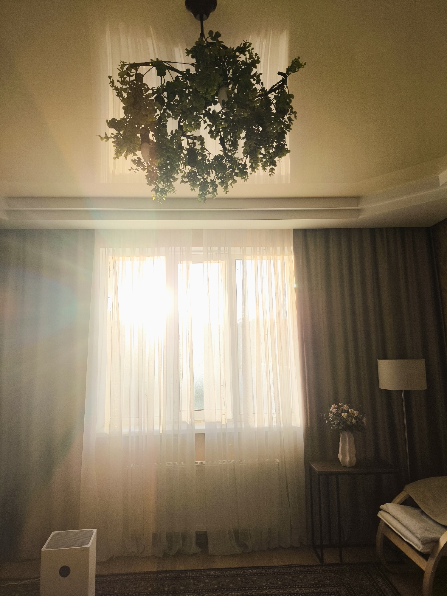 The image shows an indoor scene, possibly a living room or bedroom, bathed in warm, natural light filtering through sheer curtains. A large window is visible, with sunlight streaming through, creating lens flares and a bright glow. A chandelier decorated with greenery hangs from the ceiling. To the right, a tall floor lamp stands next to a chair with cushions. A small side table holds a vase with a bouquet of flowers. On the left side of the image, a white air purifier is placed on the floor. The room has dark curtains drawn on one side and a patterned rug on the floor. The overall atmosphere is serene and well-lit.