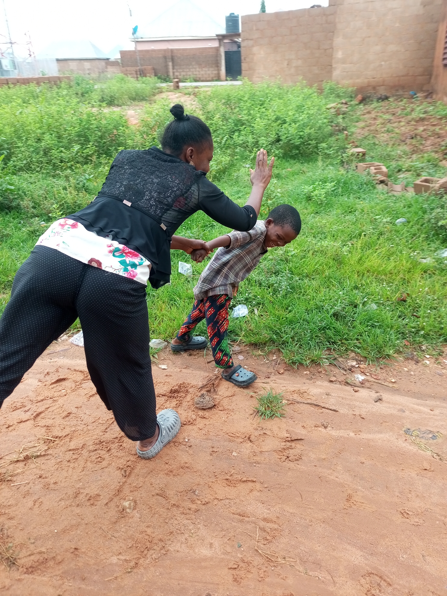 In Tempure, Nigeria, a young boy with dark skin and short hair is engaged in an interaction with an adult woman. The boy, wearing a plaid shirt and colorful patterned pants with black Crocs, appears to be laughing or shouting, with his head tilted back and eyes closed. He is in a bent-over position, with his right arm extended and hand open. The woman, with her dark hair in a bun, wears a black lacy top and black polka-dot pants with a floral-patterned belt. She is holding the boy's right hand and has her left hand raised. They are positioned on a dirt path with sparse green grass and some debris, including bricks and plastic bottles, scattered in the background. Beyond the immediate area, there are buildings, including one made of reddish-brown brick and another with a water tank.