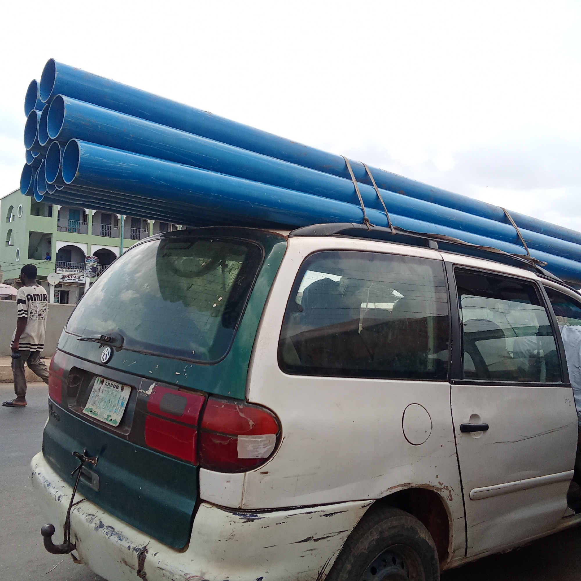 A white and green minivan, with visible damage and rust, is heavily loaded with blue pipes strapped to its roof. The vehicle has a Lagos license plate. A pedestrian in a striped shirt and trousers walks past the car. In the background, a building with green and white paint is visible. This is in Lagos, Nigeria.
