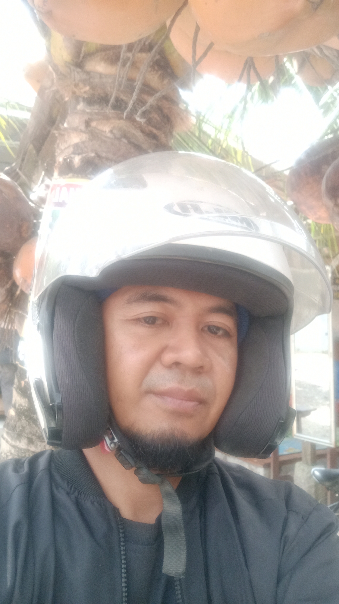 An outdoor, medium-shot, selfie of an adult male wearing a white and clear-visored motorcycle helmet and a dark blue zip-up jacket over a dark shirt. The man has light brown skin, a goatee, and appears to be looking forward or slightly to the right. The helmet covers his hair and ears, with the chin strap visible and buckled. The background is blurry, showing what appears to be a palm tree with coconuts hanging above and some greenery. The image has a bright, blown-out sky. The scene suggests a location in Cibeber, Indonesia, possibly a rural or tropical area due to the presence of the palm tree and coconuts. The lighting indicates it is daytime, likely sunny. There are no other people clearly visible in the frame, and no discernible action or interaction is taking place. The overall impression is casual and informal.