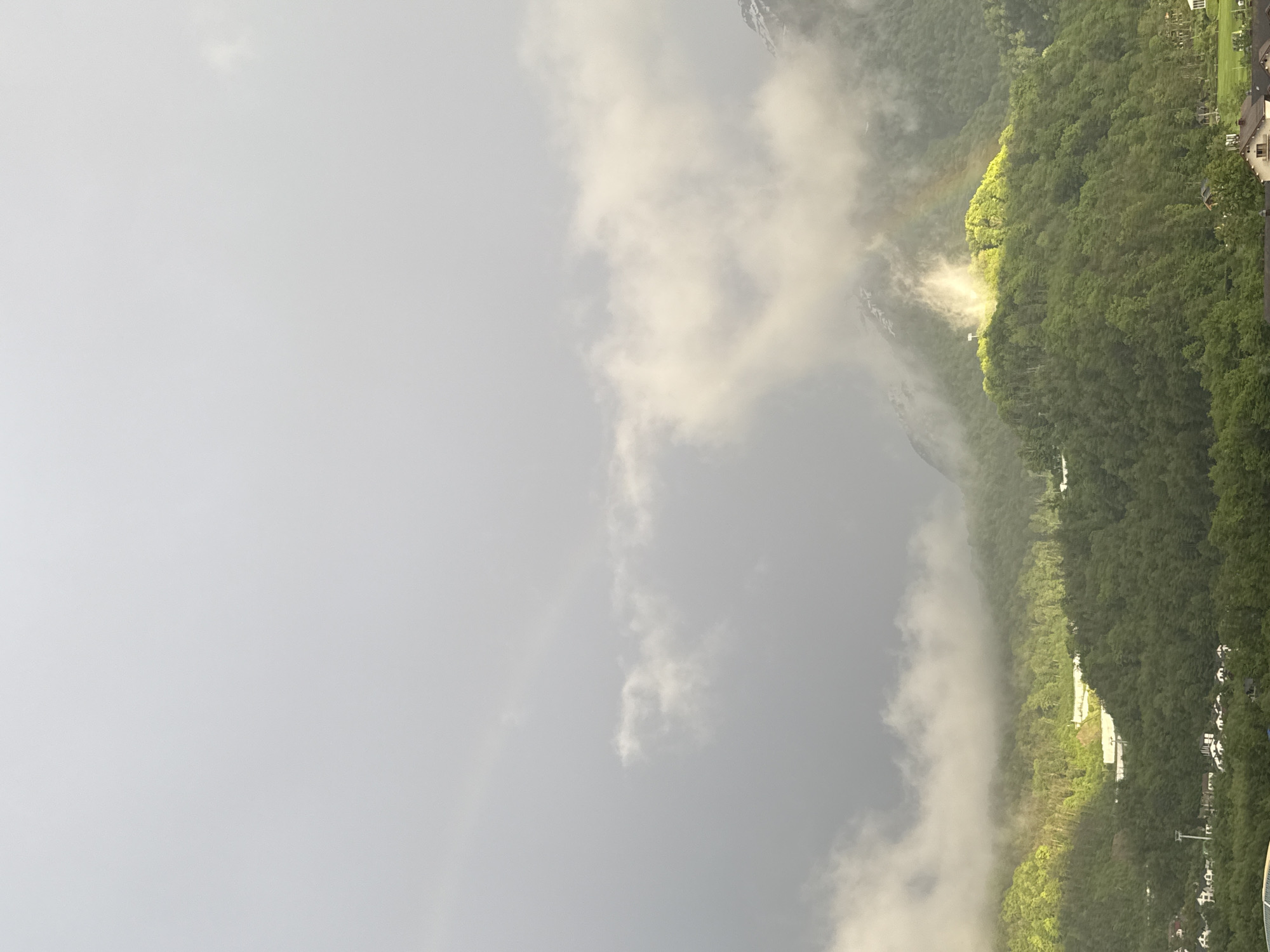The image shows a mountain landscape partially obscured by fog, with a faint rainbow visible in the lower left corner. The right side of the image reveals a forested mountainside with some buildings, potentially residential, visible at the base. The location appears to be Krasnaya Polyana, Russia, based on the typical landscape of the area. The sky is overcast and gray. The lighting suggests that the sun is shining behind the clouds or fog, illuminating parts of the mountainside.