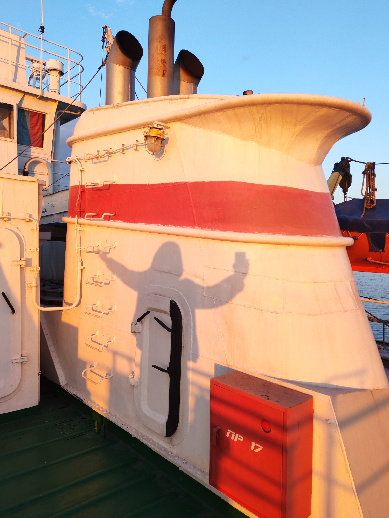 The image shows a portion of a white ship with a red stripe, including its smokestacks and a section of the deck, under warm lighting, likely at sunset or sunrise. A shadow resembling a person holding a smartphone is cast on the side of the ship. A red box with white lettering "ПР 17" is mounted on the side. The location is in the city of Unknown, Unknown.