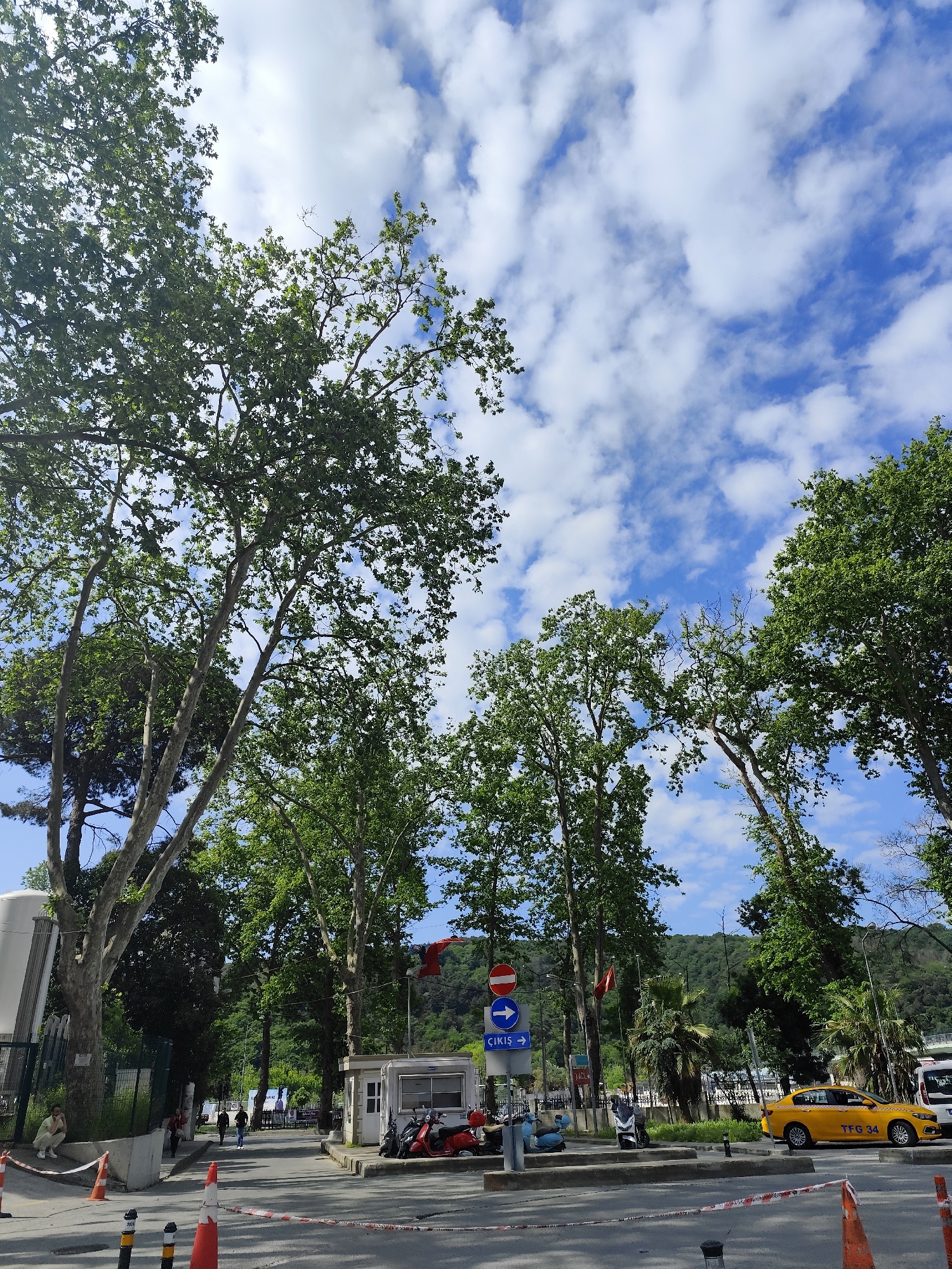 The image shows a street scene in Sarıyer, Turkey, with a view of a road lined with trees. The sky is blue with scattered clouds.
To the left, a person sits on a ledge near a fence. Several people are walking along the sidewalk in the background. On the right side of the road, a yellow taxi is parked with the license plate "TFG 34". Various motorbikes and a scooter are parked near a small building with signage.
A street sign with a red and white "do not enter" symbol, a blue arrow, and the word "Çıkış" is visible. Construction cones and barrier tape are placed in the foreground, suggesting ongoing construction or temporary street closure. A Turkish flag is visible in the background.