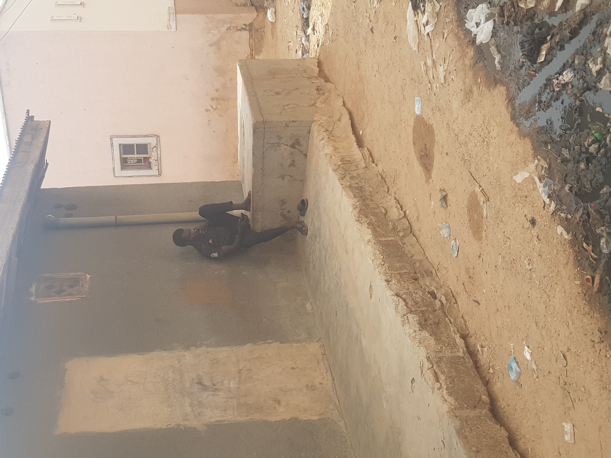 The image shows a man lying on the ground next to a concrete structure in an outdoor area. The location is in Dutsinma B, Nigeria. The surrounding environment includes buildings with visible windows, a wall with a pipe running along it, and a dirt-covered area with debris and what appears to be a ditch filled with water and refuse.