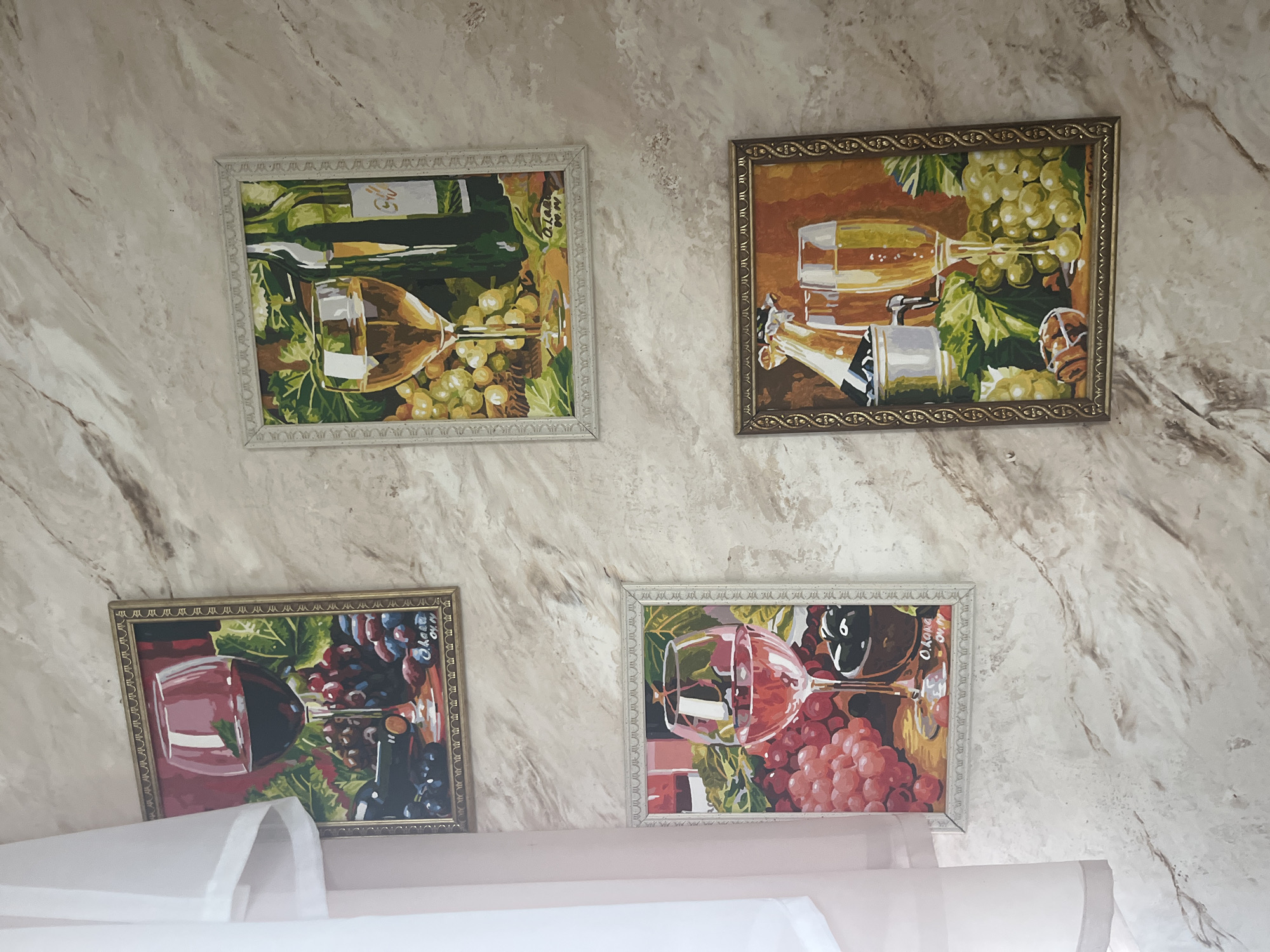 Four framed paintings of wine and grapes are displayed on a wall with a marble-like pattern. The paintings are arranged in a two-by-two grid, with the top left and bottom left paintings being taller than the top right and bottom right paintings. The top left painting depicts a glass of red wine, a bottle, and dark grapes. The top right painting features a glass of white wine and a green bottle with white grapes. The bottom left painting shows a glass of rosé wine with red grapes and olives. The bottom right painting illustrates two glasses of champagne, a bottle in a cooler, and green grapes. The wall is a neutral beige color with brown and white marbling. A white sheer curtain is visible on the left side of the image. There are no people or animals in the scene. The location is described as Vladimirovka, Russia.