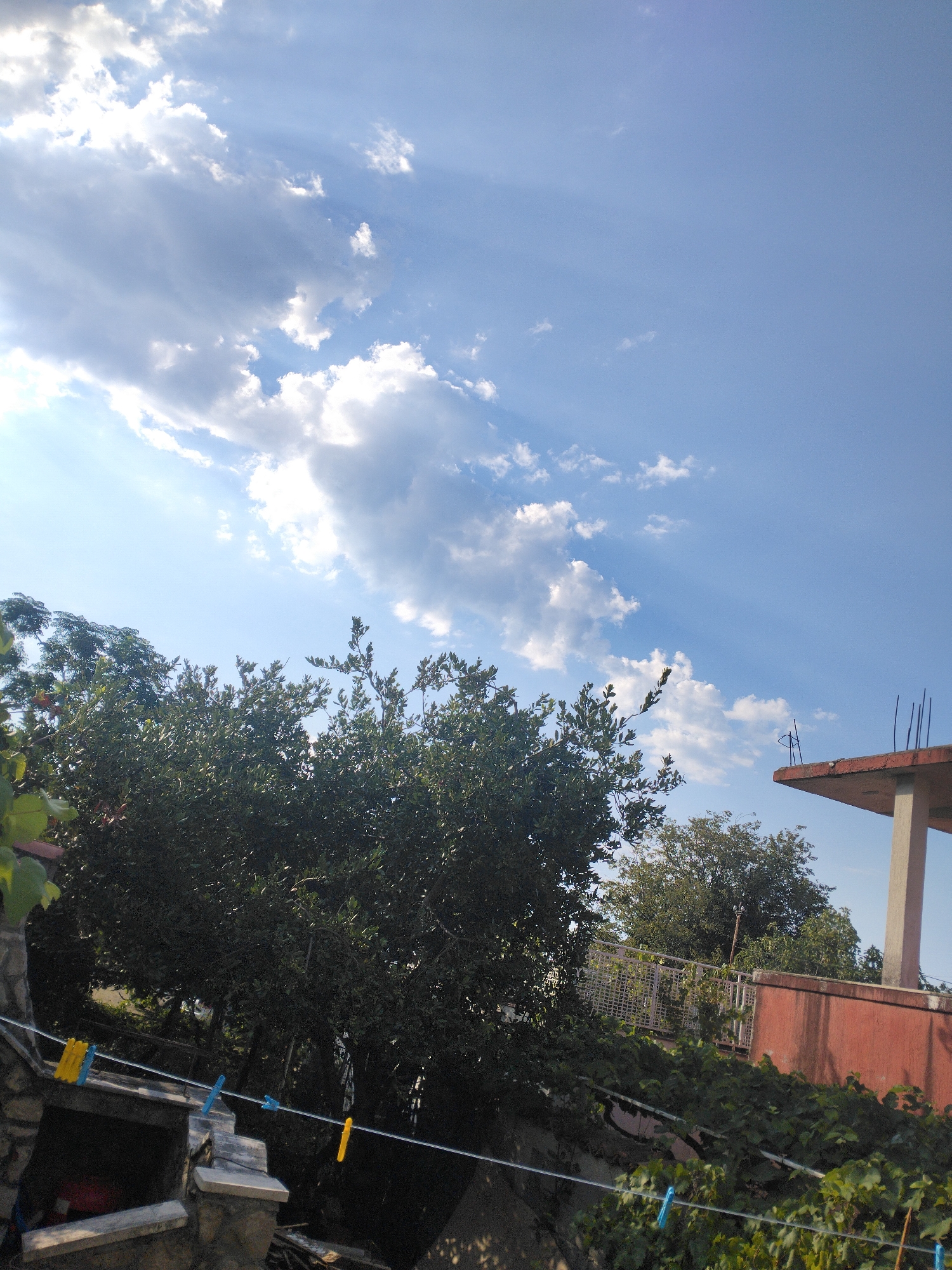 The image captures a view of the sky and vegetation from an elevated vantage point, likely a balcony or rooftop, in Zgrade, Montenegro. The sky is a clear blue, with scattered white clouds that are illuminated by visible sunbeams streaming through them. In the foreground, dense green foliage from trees dominates the lower portion of the frame. To the right, a portion of a building is visible, featuring a reddish-orange wall and a concrete support column. Clotheslines with several colorful clothespins are strung across the scene, suggesting a residential area. The overall impression is one of a sunny day in a somewhat rural or suburban setting.
