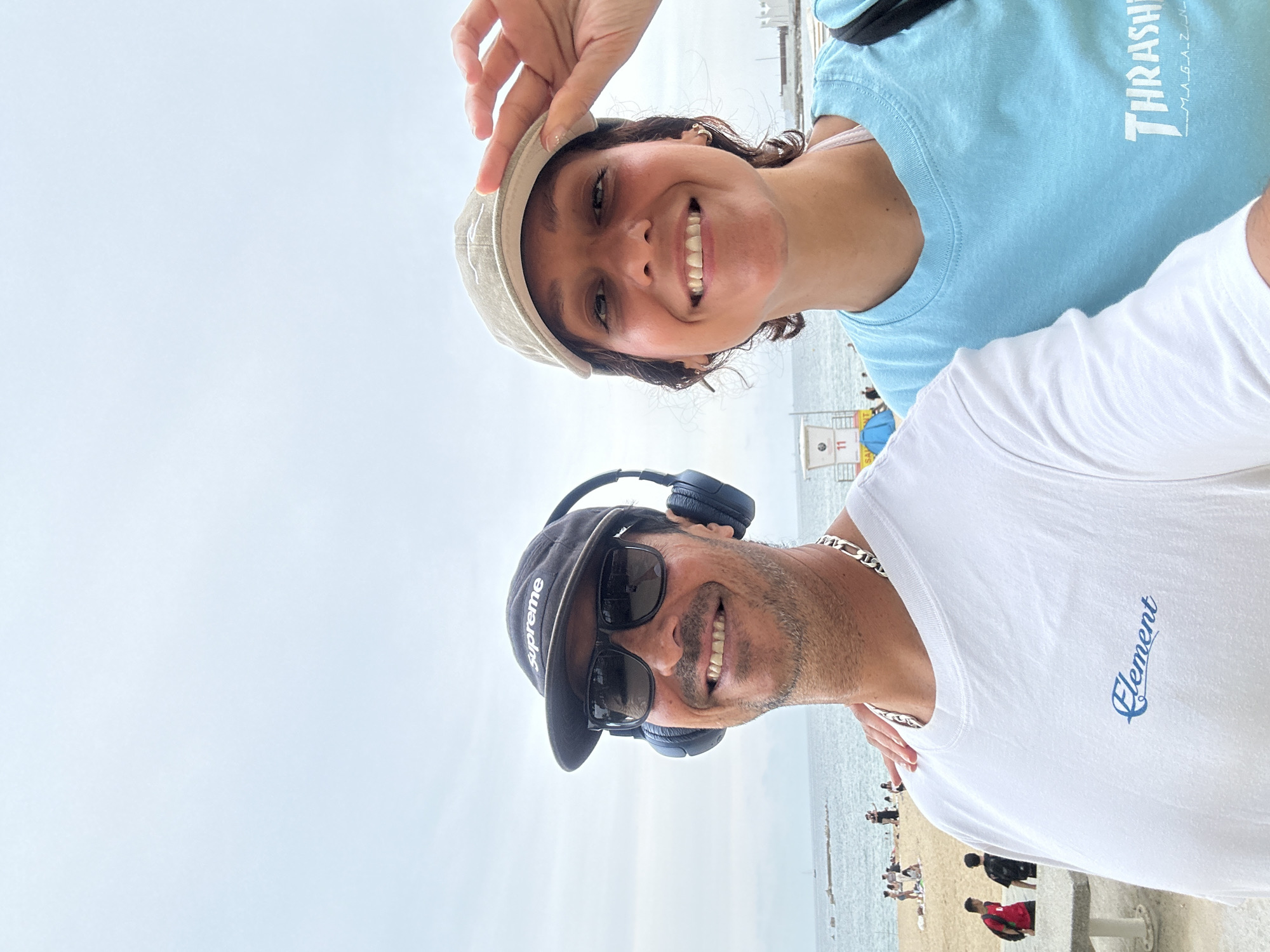 Two people pose for a selfie on Barceloneta Beach in Barcelona, Spain. The woman on the top is fair-skinned and is wearing a light beige cap, a light blue t-shirt with a white logo on it, and small gold earrings. She is holding the edge of her cap. The man below her is wearing a black cap, black sunglasses, and blue headphones around his neck. He is wearing a white t-shirt with the word "Element" written on it in cursive. A silver chain necklace is visible around his neck. They are both smiling, and the beach is visible in the background. People are walking on the beach behind them. The sky is cloudy.