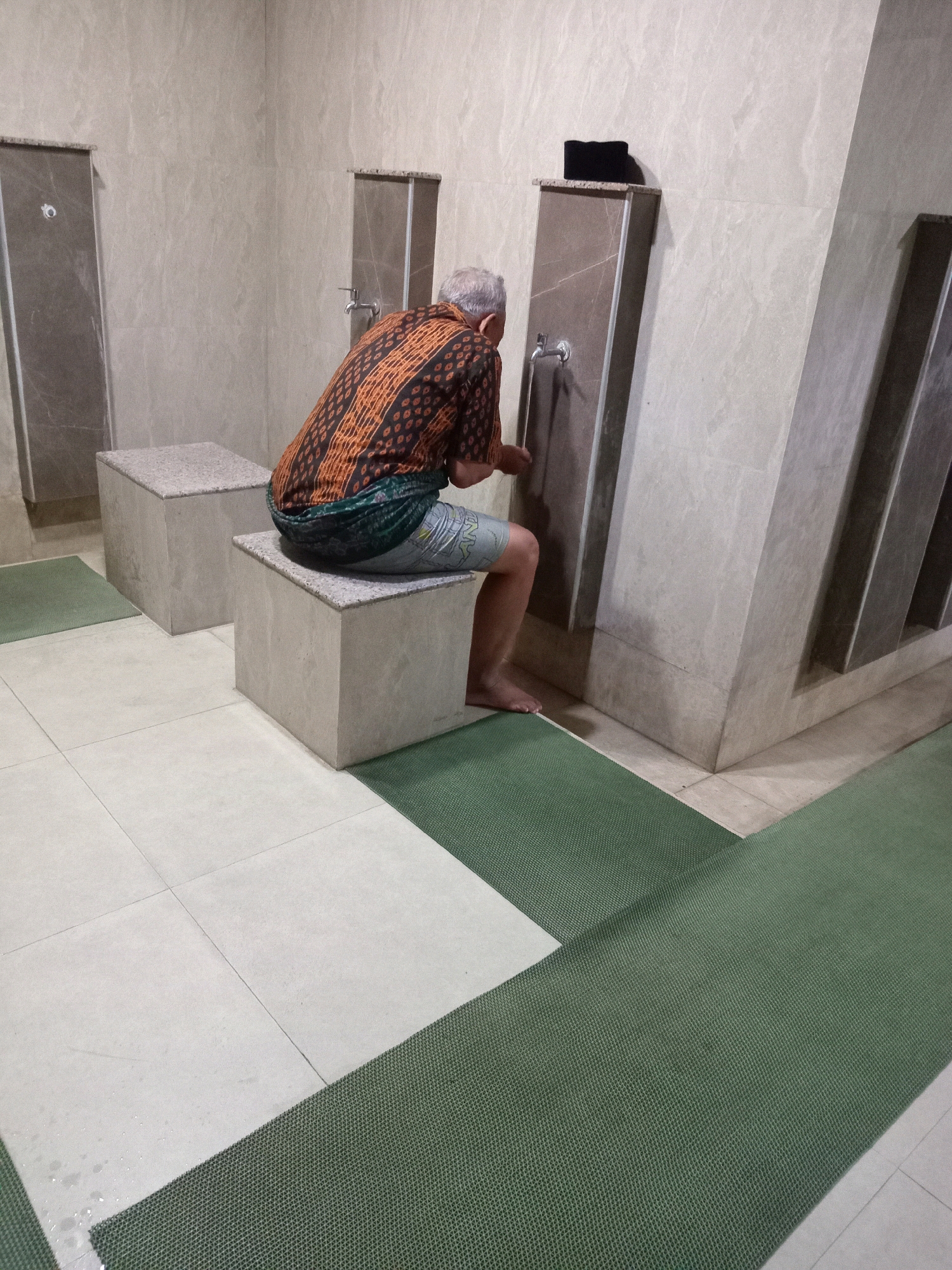 An older man with gray hair is performing ablution (Wudu) at a public facility in Randuagung, Indonesia. He is seated on a square, grey stone bench, facing a wall with multiple spigots. The man is wearing a patterned brown and orange shirt and a green sarong wrapped around his waist. He is leaning forward, washing his hands under one of the spigots. The environment appears to be a clean, tiled area, likely part of a mosque or public restroom, with large, light-colored tiles on the walls and floor. Green mats are laid out on the floor in front of the ablution stations. A black peci (cap) rests on a marble-like surface above the man. The lighting is somewhat dim.