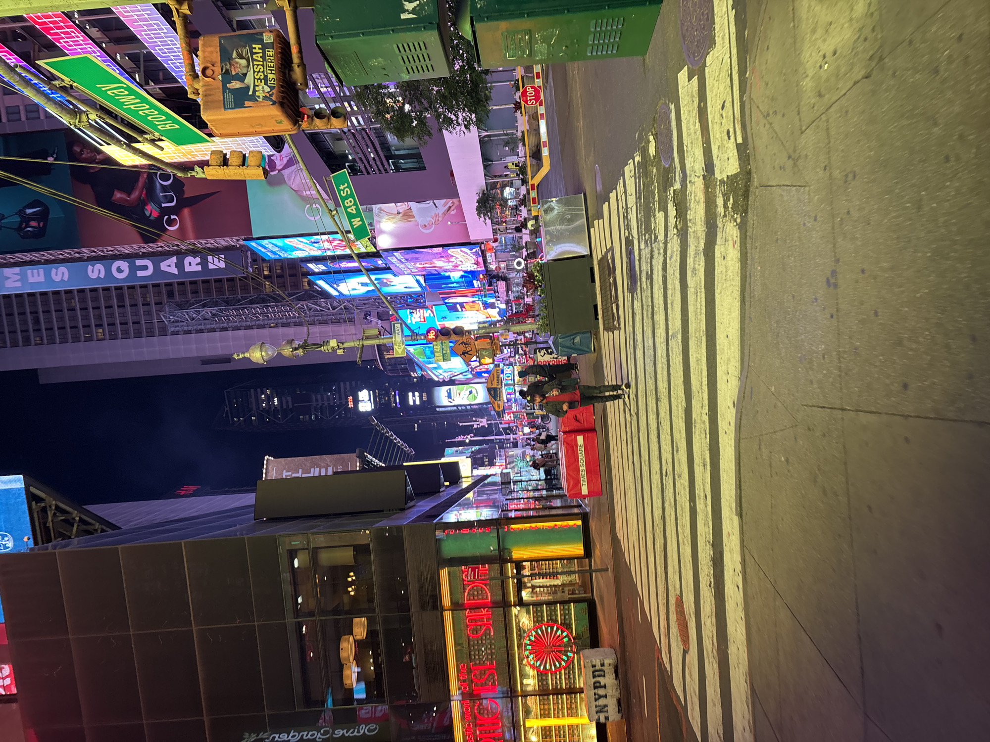 The image captures a vibrant nighttime street scene in Times Square, New York City, looking down a thoroughfare brightly lit by countless electronic billboards and neon signs. The time of day is clearly night, as indicated by the dark sky overhead and the dominance of artificial light illuminating the entire area. The ground appears slightly wet, reflecting the colorful lights from the surrounding advertisements.
Several pedestrians are visible, with two individuals in the mid-ground walking on the crosswalk and others in the distance, engaging in typical city sidewalk activity. The street itself has a prominent crosswalk marked with yellow lines, reflecting the bright lights.
Key location cues confirm the setting as Times Square, New York, including street signs for "Broadway" and "W 48 St," and explicit text visible on a red box that reads "TIMES SQUARE" and a white block displaying "NYPD." A "STOP" sign is also visible. Large advertisements dominate the visual landscape, featuring various brands and messages. Visible text on these signs includes fragments like "MES SQUARE," "GUC" (likely part of Gucci), and a prominent sign reading "MESSIAH IS HERE!" Other less clear texts include "stic work of the," "2026," "Lou Walters," "OR," and "BRETT." The overall atmosphere is bustling and commercial, characteristic of this iconic New York landmark.