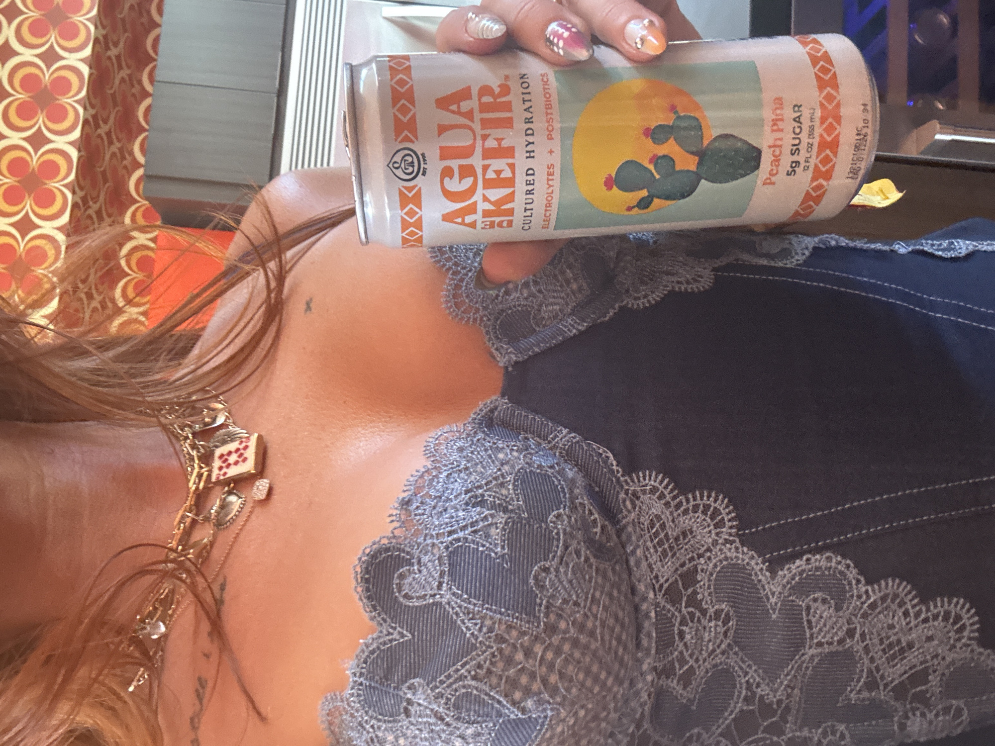 The image shows a woman holding a can of Agua Kefir Peach Piña. She is wearing a blue top with lace detailing, and a gold necklace with charms. Her nails are decorated with colorful designs. The background includes a patterned curtain and what appears to be part of a piece of furniture. There is no indication that this image was taken in the city of Indio, United States of America.