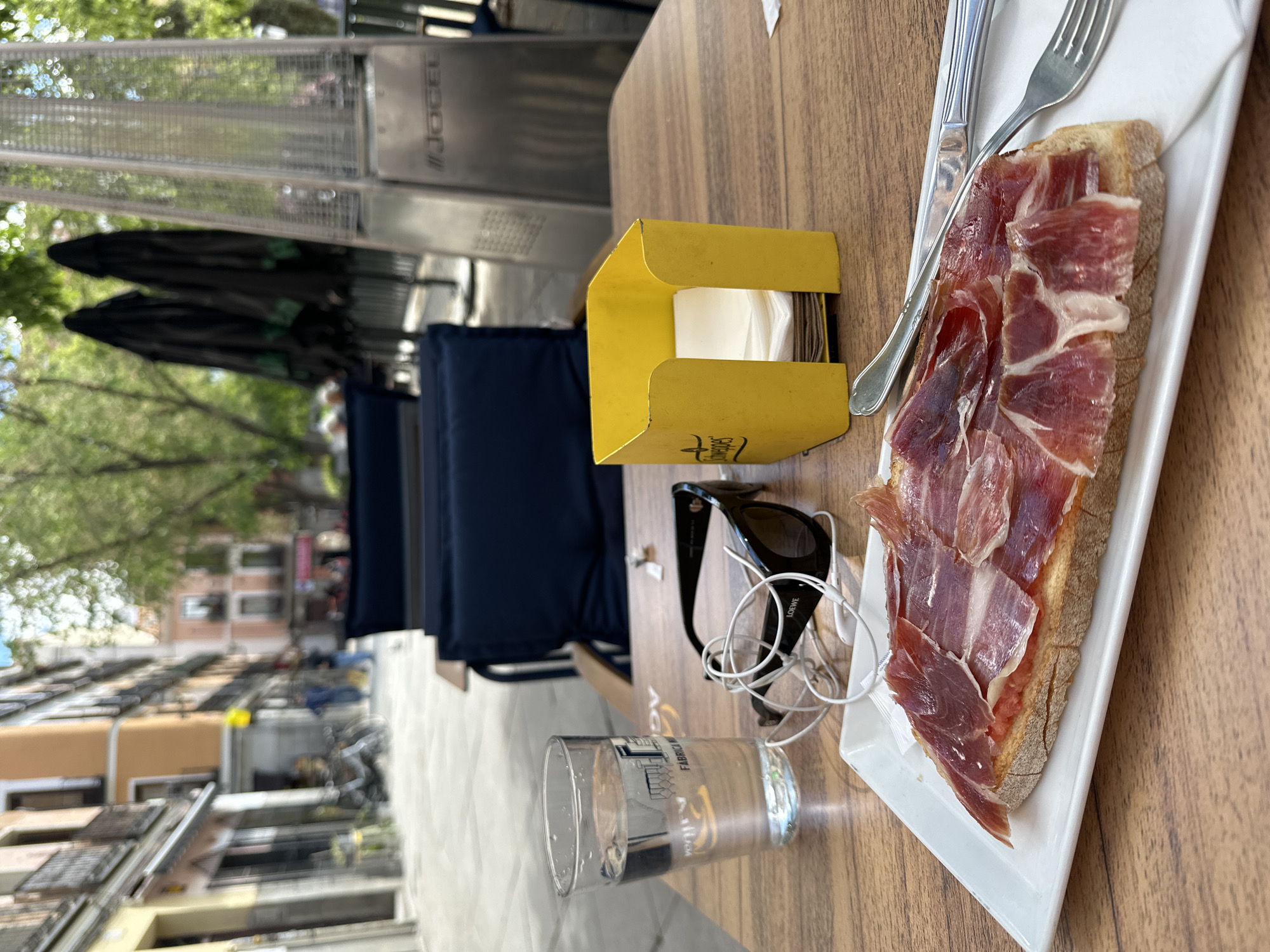 The image shows a table at an outdoor café, likely in Madrid, Spain, based on the architectural style visible in the background. On the table, there's a plate with a slice of bread topped with "jamón ibérico," a knife and fork, a yellow napkin holder with napkins, a glass of water, a pair of sunglasses, and a set of earphones. The table is wooden and reflects outdoor light. Behind the table are outdoor seating cushions and heating lamps, suggesting a shaded outdoor area. In the background is a street with residential buildings.