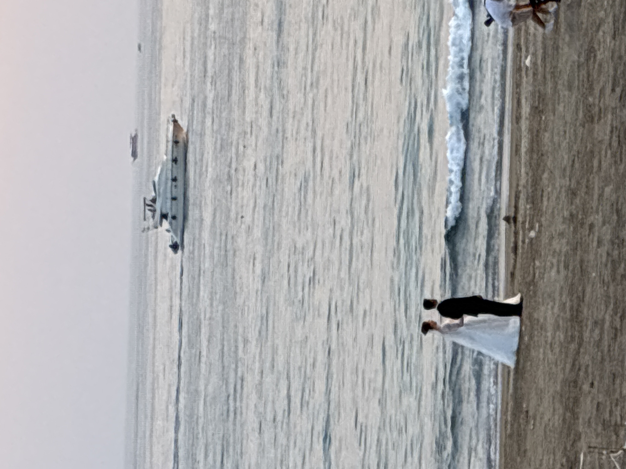 A wedding couple, a bride in a white gown and a groom in a black suit, stand on a sandy beach facing the sea. The bride is to the left of the groom, and they are looking out at the water. In the background, a large white yacht sails on the ocean, with several smaller boats visible further out. The sky is overcast and the sea is a muted gray-blue. The beach appears to be located in Alanya, Turkey, judging by the characteristic style of yachts and the general coastal landscape.