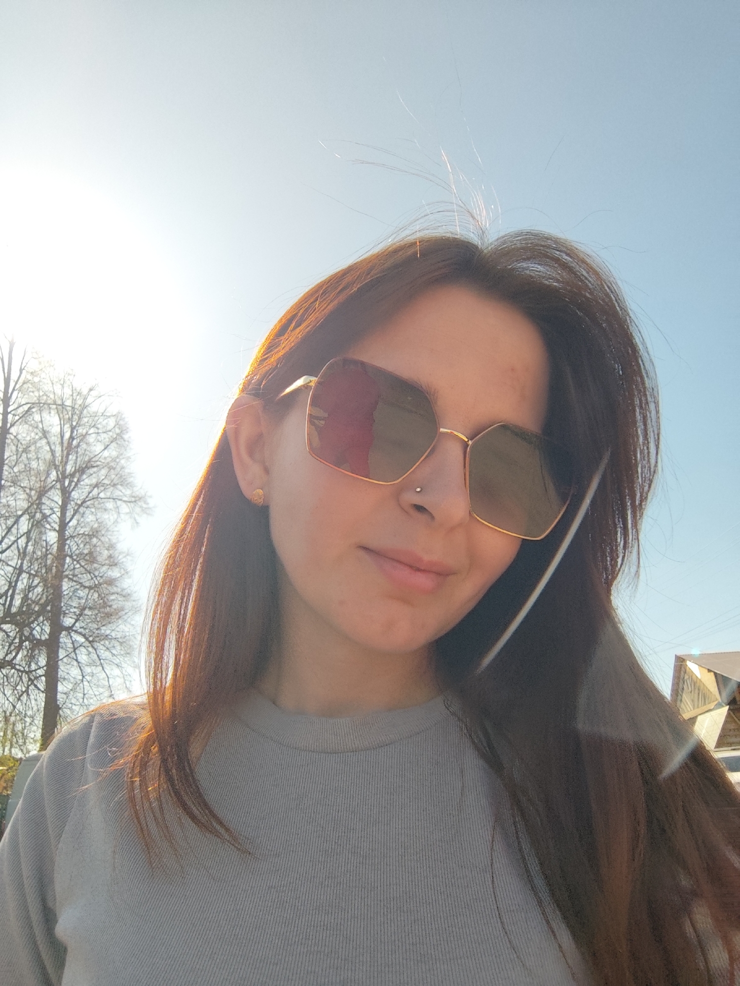 A woman with light brown hair and sunglasses is centered in the image. She is wearing a gray top. She has a nose piercing and earrings. A tree and a building are in the background. The sky is blue. The picture was taken outdoors in daytime, possibly in bright sunlight. The exact location in the city of Фоки, Russia cannot be determined from the image.