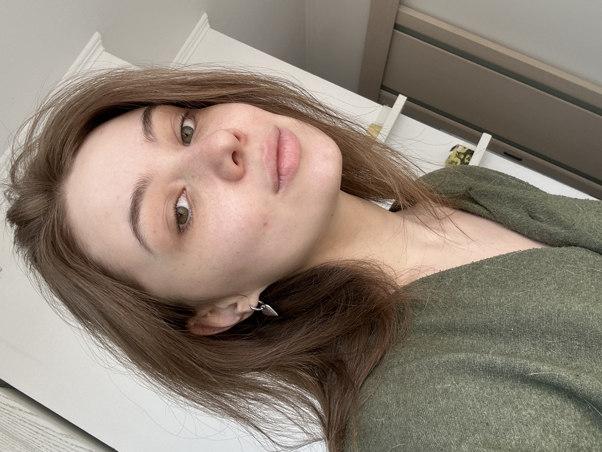 A young woman with light skin and brown hair, wearing a green sweater and a silver earring, is the primary subject of the image. Her gaze is directed towards the viewer. The background includes a white door and part of a brown sliding door or window frame. The location cannot be determined based on the visual information provided.