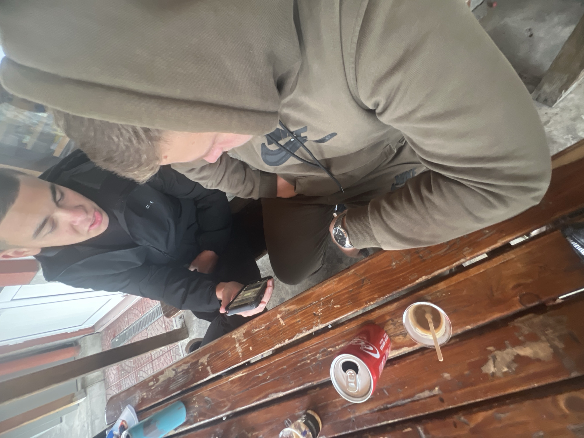 Two individuals are seated at a rustic wooden table. One person, wearing a black jacket, is holding a smartphone. The other, wearing a khaki-colored Nike hoodie with the hood up, is also seated at the table. On the table are a can of Coca-Cola, a drink in a clear plastic cup with a stirring stick, and what appears to be another beverage container. The scene is situated in Asenovgrad, Bulgaria, as indicated by geotags. The environment suggests an outdoor setting, potentially a cafe or patio, based on the table and the visible exterior elements. The people appear to be engaged in a casual activity, possibly social interaction or using their phones.