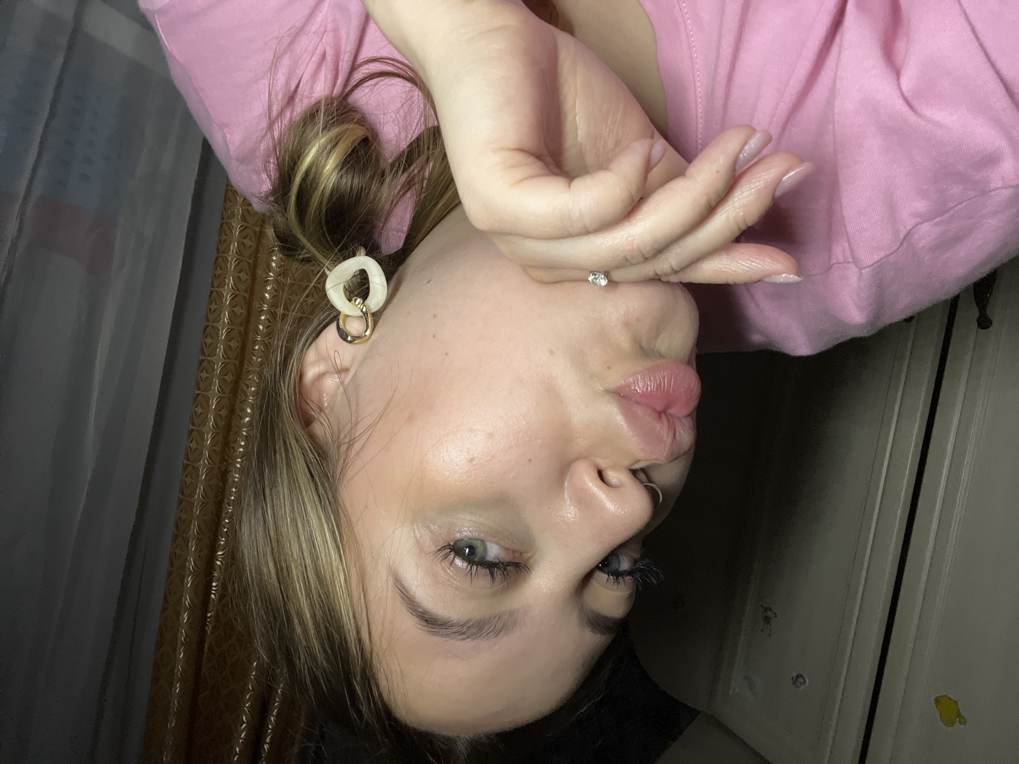A person is shown in a close-up shot, oriented upside down. The person has light skin, light brown hair with blonde highlights pulled back in a bun, and is wearing a pink top. They have earrings in both ears, one featuring a white and gold linked design, and a stud in the other. Their hand is partially covering their face, and they appear to be pouting slightly. A septum piercing is visible on their nose. The background includes a curtain on the left and what appears to be a door on the right. The location is not apparent from the image.