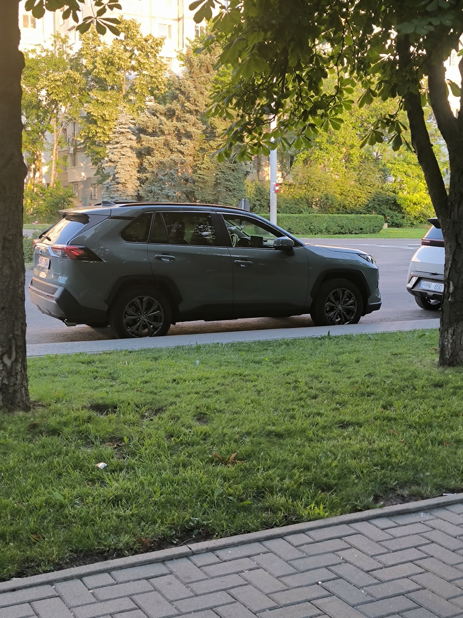 A dark green Toyota RAV4 is parked on the side of a street in Chișinău, Moldova, with its front right tire slightly deflated and resting on the curb. The car is viewed from its left side. In the background, a white car with a license plate reading "YHW 650" is also parked on the street. Behind the cars, there is a manicured hedge and trees, with a building visible through the foliage. The foreground shows a paved walkway and a grassy area with some scattered leaves and what appears to be a cigarette butt. The scene is illuminated by the golden light of late afternoon or early morning sun, casting long shadows.