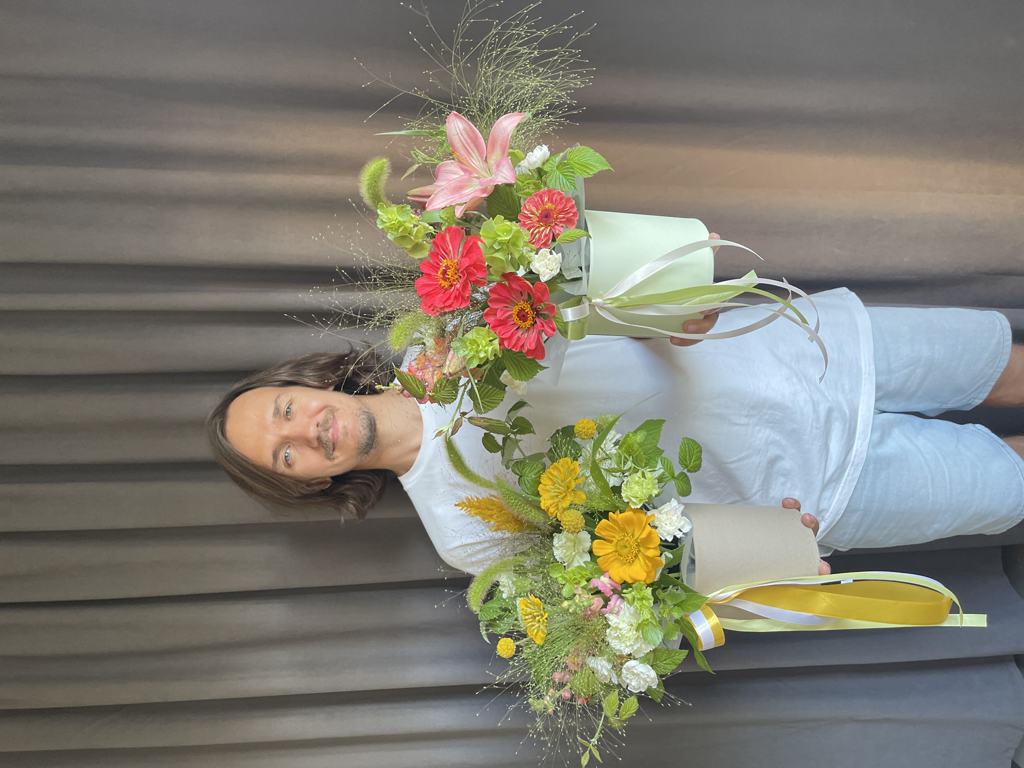 A man with medium-length brown hair and blue eyes stands against a gray curtain backdrop, holding two floral arrangements. The arrangement on his left features yellow and white flowers, accented with greenery and yellow and white ribbons. The arrangement on his right contains pink lilies and red zinnias, also with greenery and light green and white ribbons. The man is wearing a white t-shirt and light blue shorts. The scene appears to be indoors, possibly a studio or shop, in Izhevsk, Russia.