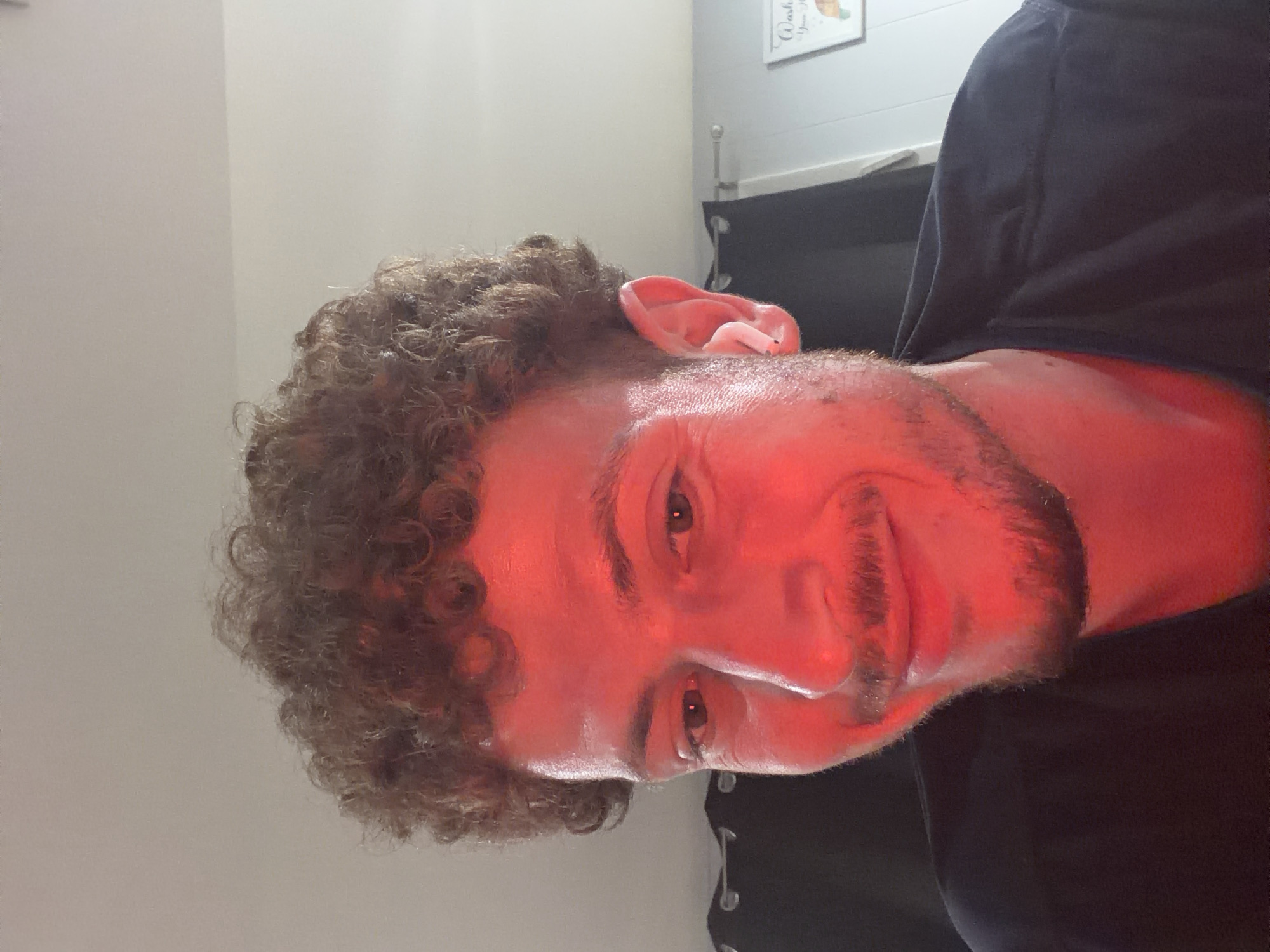 An indoor, close-up photograph depicts a male individual with curly brown hair and a light beard and mustache, illuminated predominantly by a strong red light source. The subject is seen from the chest up, positioned slightly off-center and looking downward and to the viewer's left with a subtle smile. His skin, particularly on his face and neck, is heavily saturated with red light, while his eyes are open and reflect small light sources. An earbud or small earring is visible in his right ear. He is wearing a dark, possibly black or navy, collared shirt. The background features a dark curtain with silver grommets on the left, a light-colored wall on the right, and a white or off-white ceiling above. A partial framed item, possibly a sign, is visible on the wall to the far right, with the words "Wash Your..." and a graphic of a hand discernible. The scene is located in Pepper Pike, United States of America.