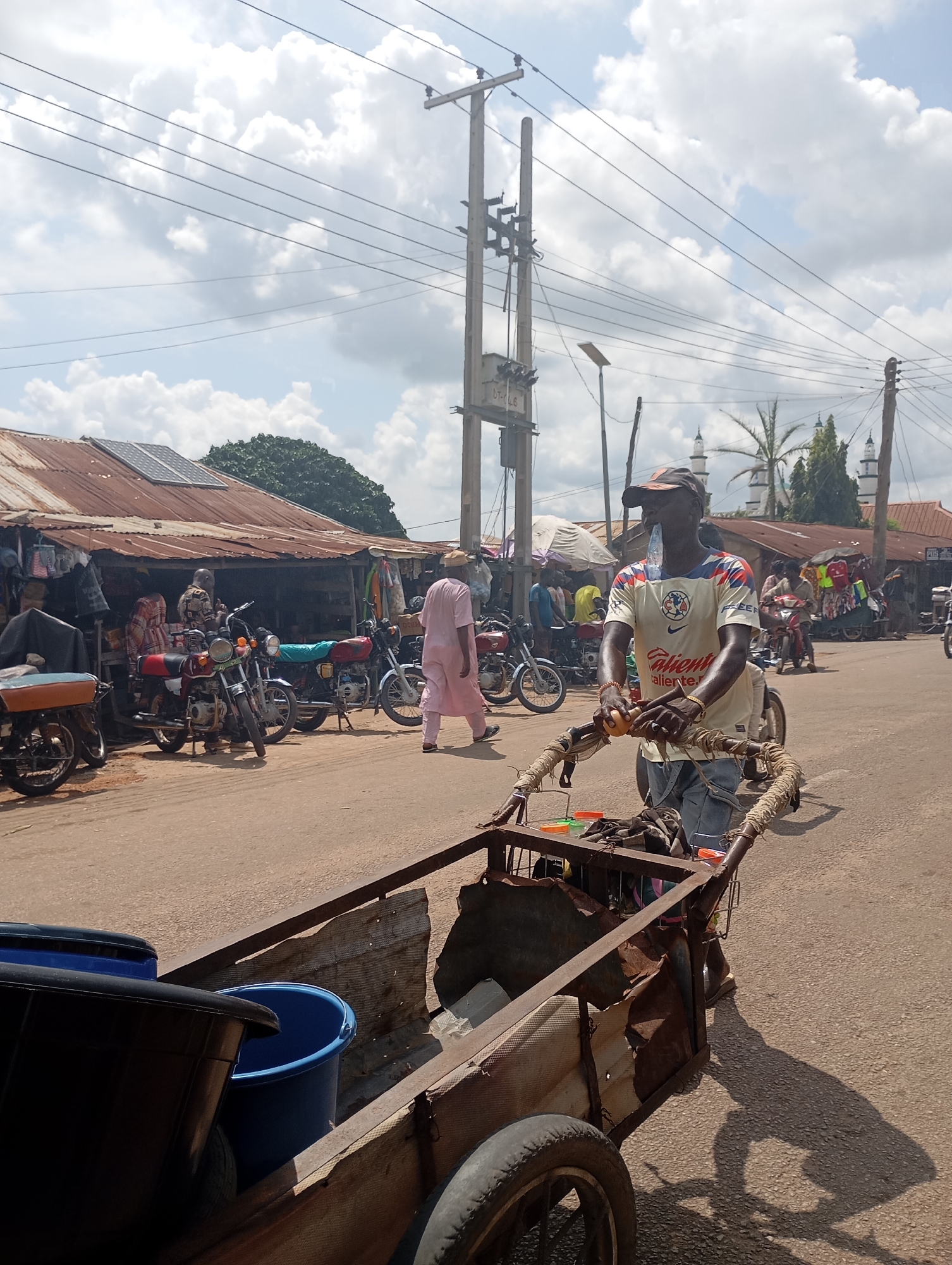 This is a full-frame, eye-level, outdoor, street-level photograph taken in Shendam, Nigeria. In the foreground, a man wearing a baseball cap, a white t-shirt with a yellow and blue logo, and blue jeans is pushing a rusty metal cart loaded with two large plastic tubs, one black and one blue. The cart has two wheels and appears to be used for transporting goods. The man is holding onto the handles of the cart and has a bottle of water in his mouth. The ground is paved with asphalt. In the background, there are several stalls with corrugated metal roofs, some displaying goods. Various motorcycles are parked in front of the stalls. A person in a pink outfit is walking away from the camera on the right side of the street. There are electrical poles with numerous wires running across the sky, and a cloudy sky with patches of blue. In the far background, several minarets of a mosque are visible. The overall scene depicts a busy market or street scene in an urban or semi-urban environment.
