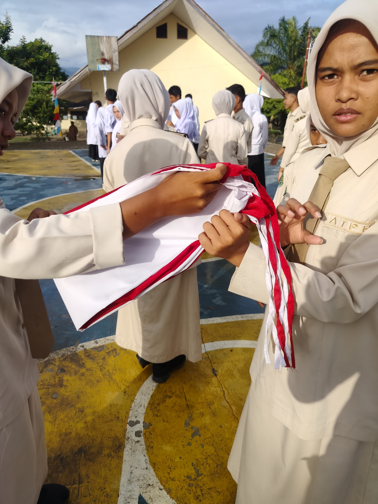 In Purwodadi, Indonesia, students in uniform are preparing to raise the national flag. The scene takes place outdoors on a basketball court, indicated by the painted lines and the hoop visible in the background. The sky is bright, suggesting it's daytime. A group of students, mostly wearing white and tan uniforms with headscarves, are gathered. Two students in the foreground are actively holding and adjusting a red and white flag. One student's arm is extended, holding a portion of the flag while the other student's hands are near the red and white stripes, seemingly preparing it for the ceremony. The students' uniforms are clean and pressed, and the flag appears to be new. The setting suggests a school event, likely a flag-raising ceremony.