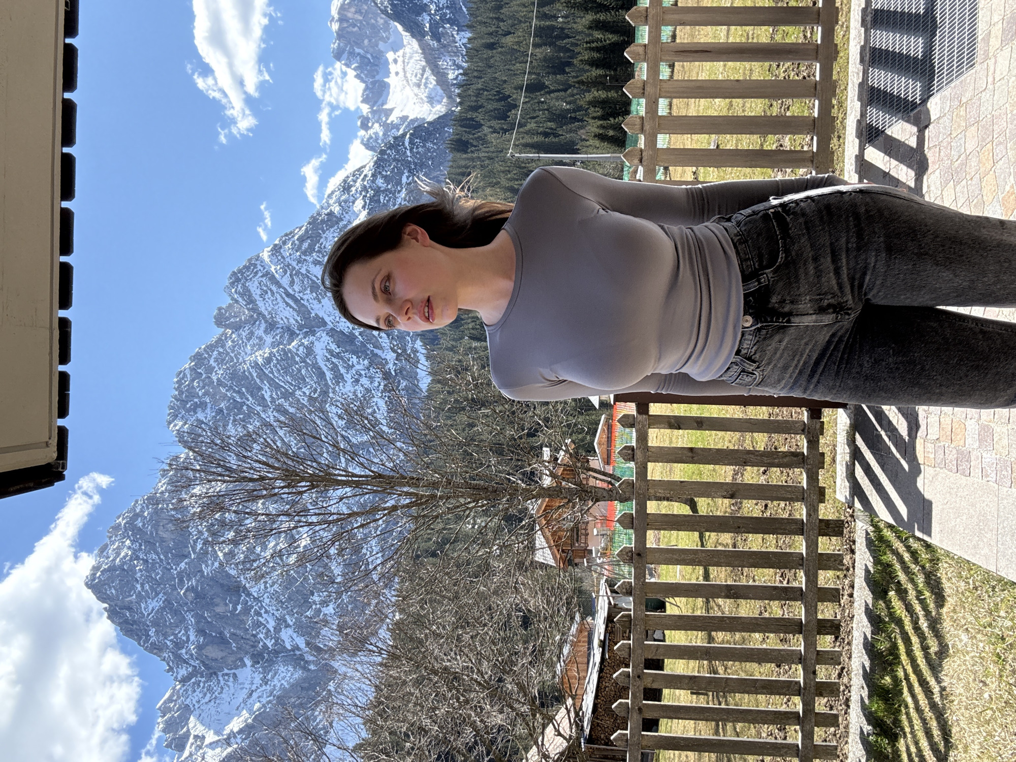 A woman stands outside in Sappada, Italy, with the Dolomites mountains in the background under a blue sky with scattered clouds. She wears a gray long-sleeved shirt and dark jeans. A wooden fence separates her from a grassy area and trees. The ground is paved. A mountain peak partially covered in snow is visible. Trees near the buildings in the village have no leaves.