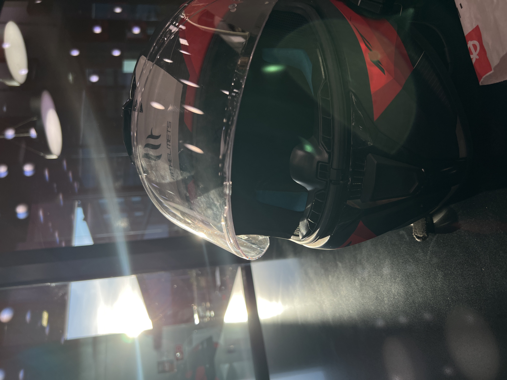 A close-up shot shows a motorcycle helmet, partially transparent, in an indoor environment, possibly a shop or display. The helmet is predominantly black with red and white accents. The visor is clear and slightly raised. The lighting suggests an interior setting, with sunlight streaming through a nearby window or glass display case. The reflection of lights and surrounding objects is visible on the helmet's surface. In the background, faint architectural details suggest an urban environment. Although not explicitly stated in the image, considering the image's source location (Sofia, Bulgaria), the scene could potentially be a motorcycle shop, an automotive accessories store, or an exhibition of safety gear.