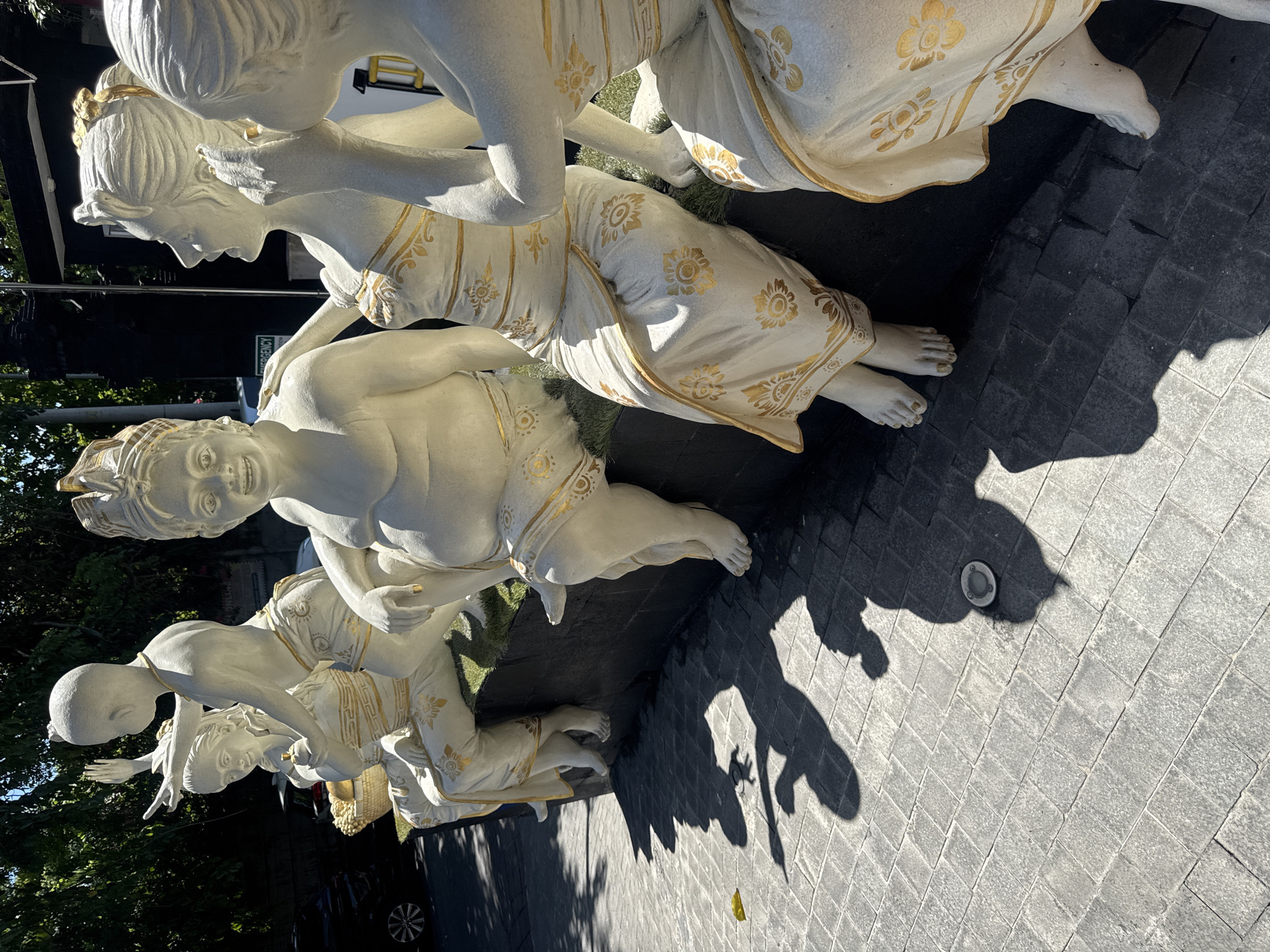 The image depicts a row of five white statues of women, likely located in Kuta, Indonesia, judging by the style of the sculptures. The statues are adorned with gold floral patterns on their garments. They are arranged in a line on a paved surface of dark grey blocks. The lighting suggests it is daytime with strong sunlight casting sharp shadows from the statues onto the pavement. The women are in varied poses; some kneeling, others standing or leaning over. One of the statues appears to be touching another's head or hair. The background includes glimpses of greenery, a vehicle, and architectural structures, suggesting an outdoor urban setting.