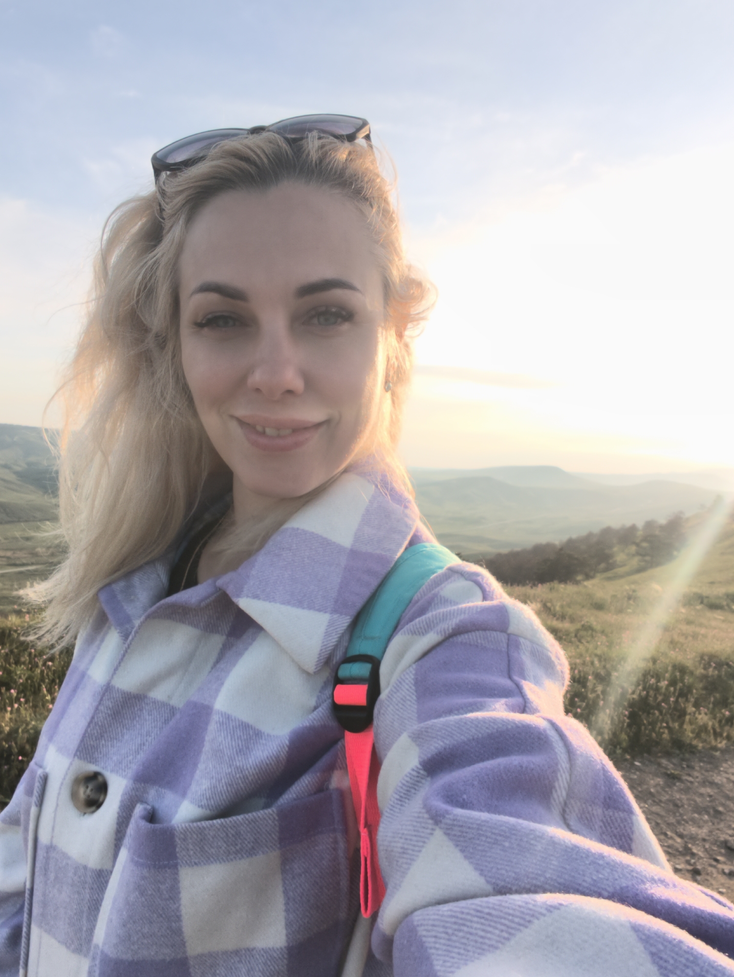 A young Caucasian woman with blonde hair and fair skin is posing for a selfie outdoors. She has sunglasses perched on her head and is wearing a lavender and white checkered shirt. A blue backpack with a pink strap is visible on her shoulder. The background shows a hilly landscape under a bright sky, possibly at sunset or sunrise. The location is Unknown, Unknown.