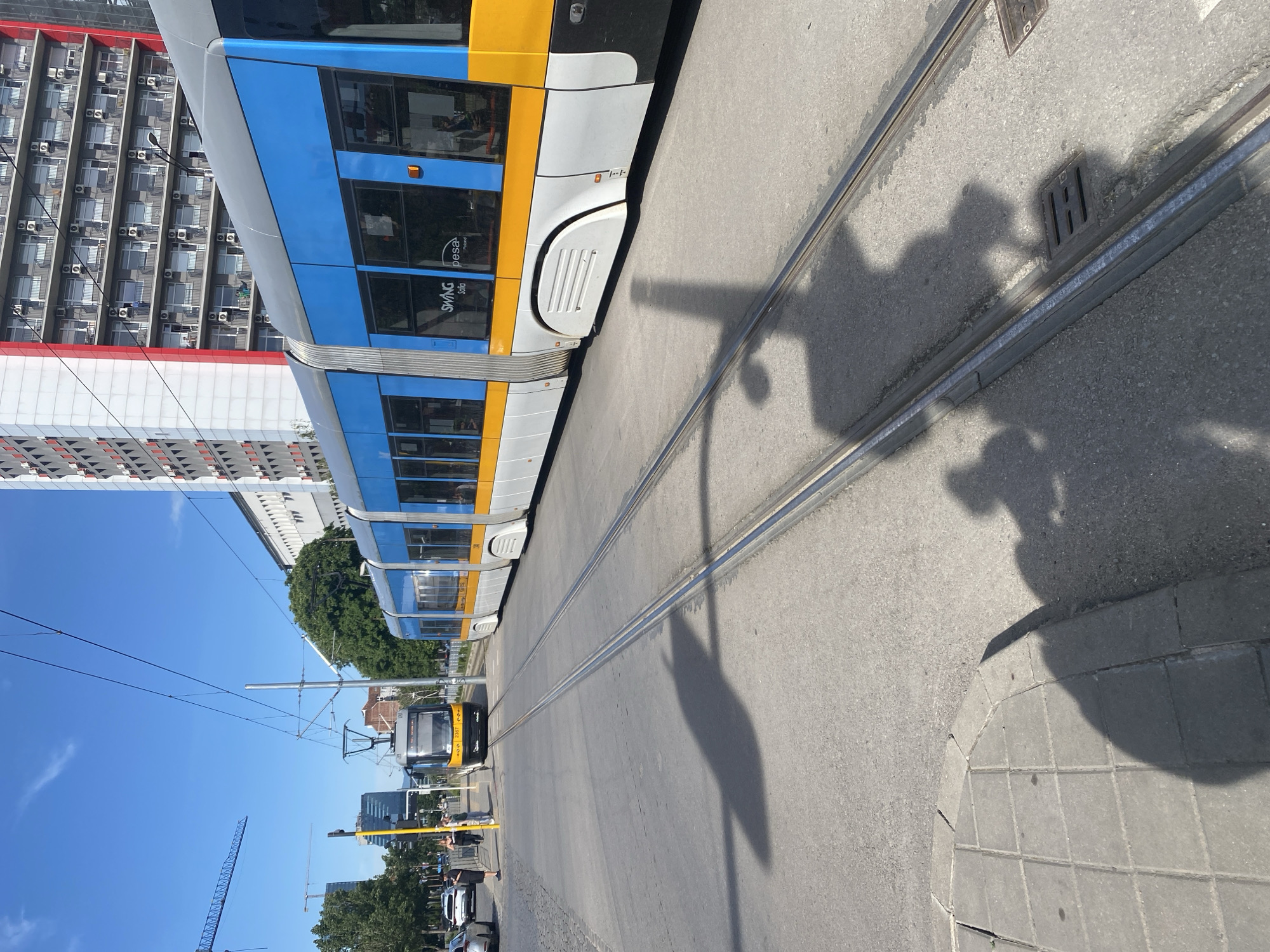 The image depicts a street scene in Sofia, Bulgaria, featuring a modern tramway system. The tram is primarily blue, white, and yellow, with the brand name "SWAG Sofia" visible on its side. Tracks run along the center of the street, with another tram visible in the distance. Buildings line the street, including a large apartment building with numerous air conditioning units visible on its façade. The sky is clear and blue. Shadows are cast on the ground, obscuring some details of the street surface. Cars and pedestrians are visible in the distance, suggesting an active urban environment. A construction crane is visible in the background, indicating ongoing development in the area.