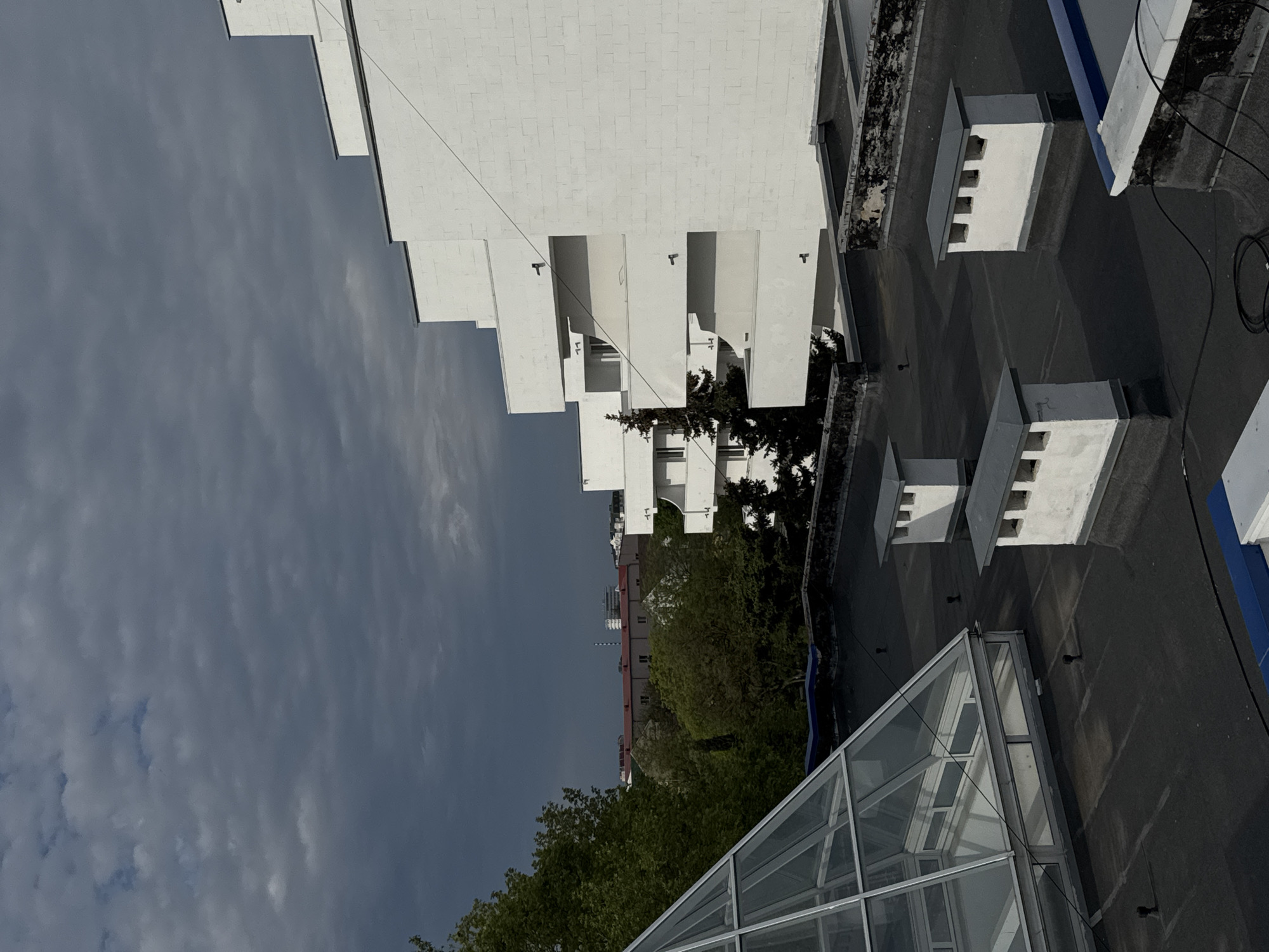 The image captures an architectural perspective of a building in Yevpatoriya, Ukraine. The structure is predominantly white, featuring a stepped design with protruding balconies or elements. The sky is overcast, with grey clouds dominating the frame. Green foliage and trees are visible, providing a natural contrast to the stark white building. A glass structure, potentially a greenhouse or skylight, is located at the lower part of the frame. It is a high-angle view. The image shows architectural details, the urban landscape, and the surrounding environment.