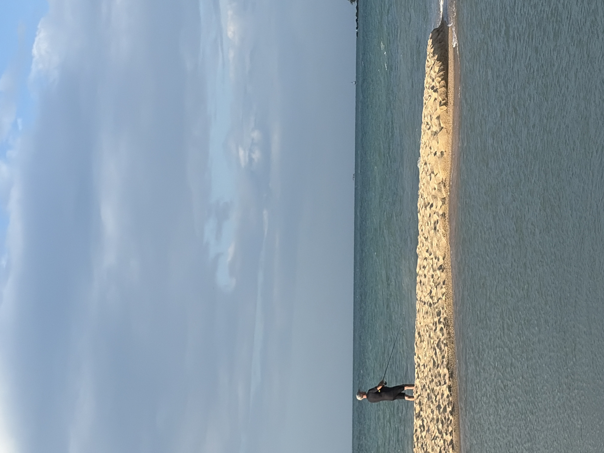 A lone person stands on a narrow strip of sandy land, fishing in the turquoise water. The person is wearing a black t-shirt and shorts, with a white cap. The sandy strip is surrounded by water, with a calm sea stretching out to the horizon. The sky above is a mix of blue and grey clouds, suggesting a partly cloudy day. The scene evokes a sense of tranquility and isolation. The specific location in Ko Samui District, Thailand is not discernible from the image.