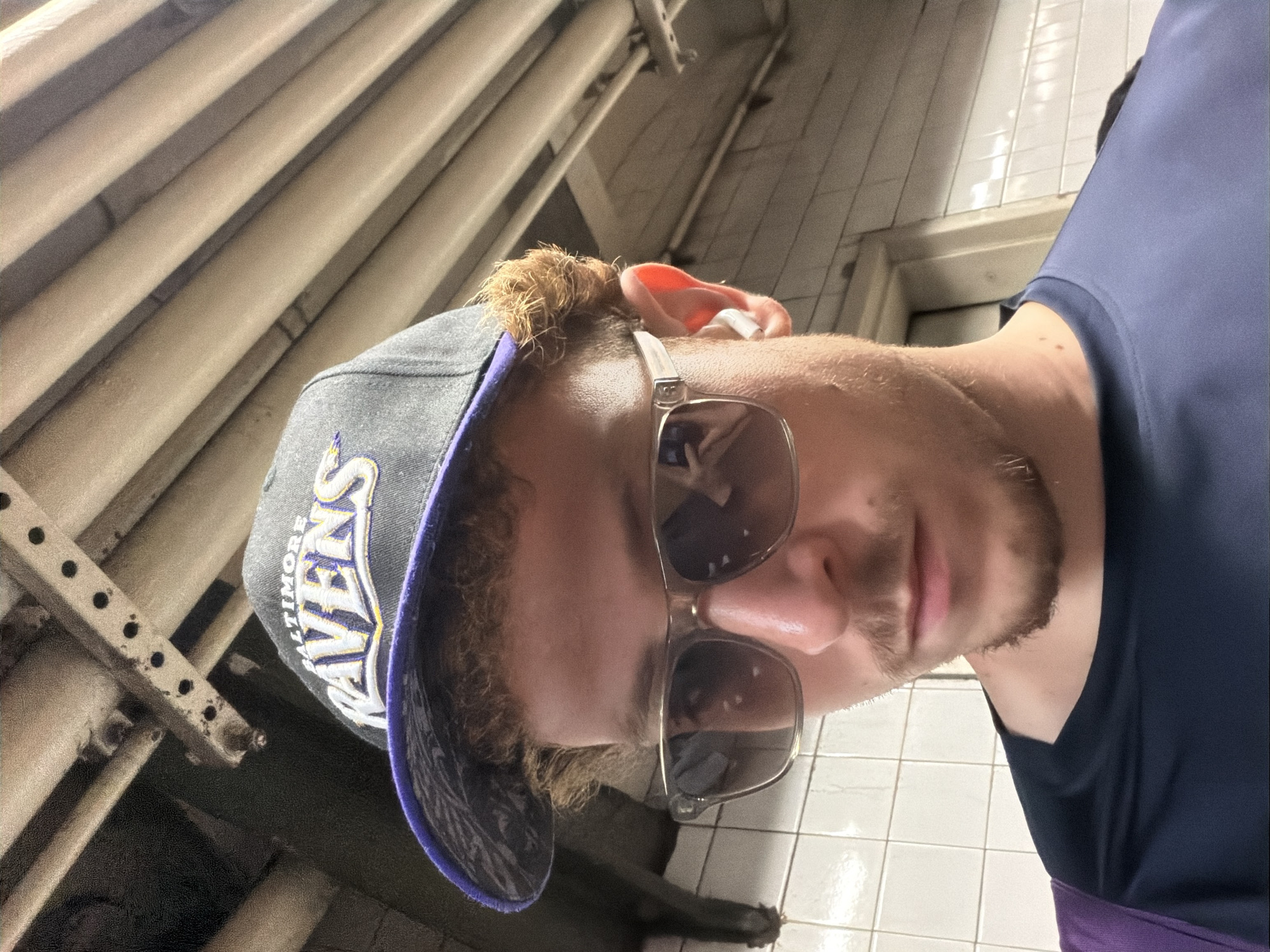 A man wearing a Baltimore Ravens baseball cap and sunglasses is standing in what appears to be a subway station in New York City. The background shows tiled walls and metal pipes, typical of the NYC subway system. The man is wearing a blue shirt.