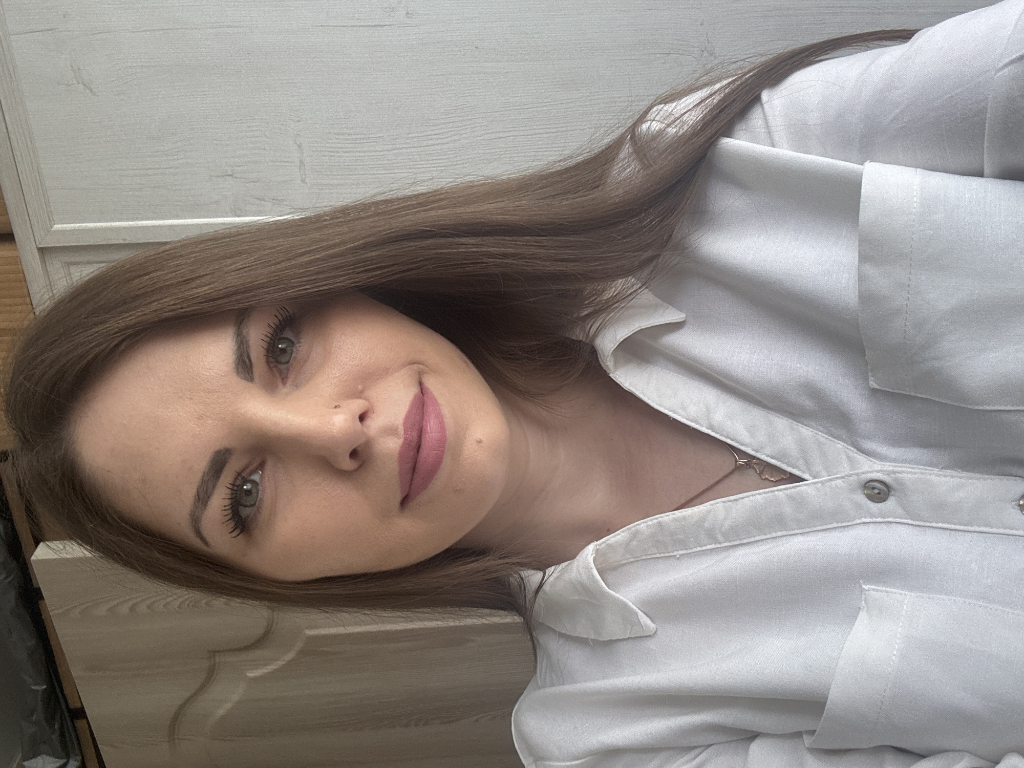 A woman with long brown hair and green eyes is wearing a white collared shirt with a pocket on the left side. She appears to be indoors in a room with light-colored wooden paneling on the wall. Based on the stated location, the image was captured in Раздолье, Russia.
