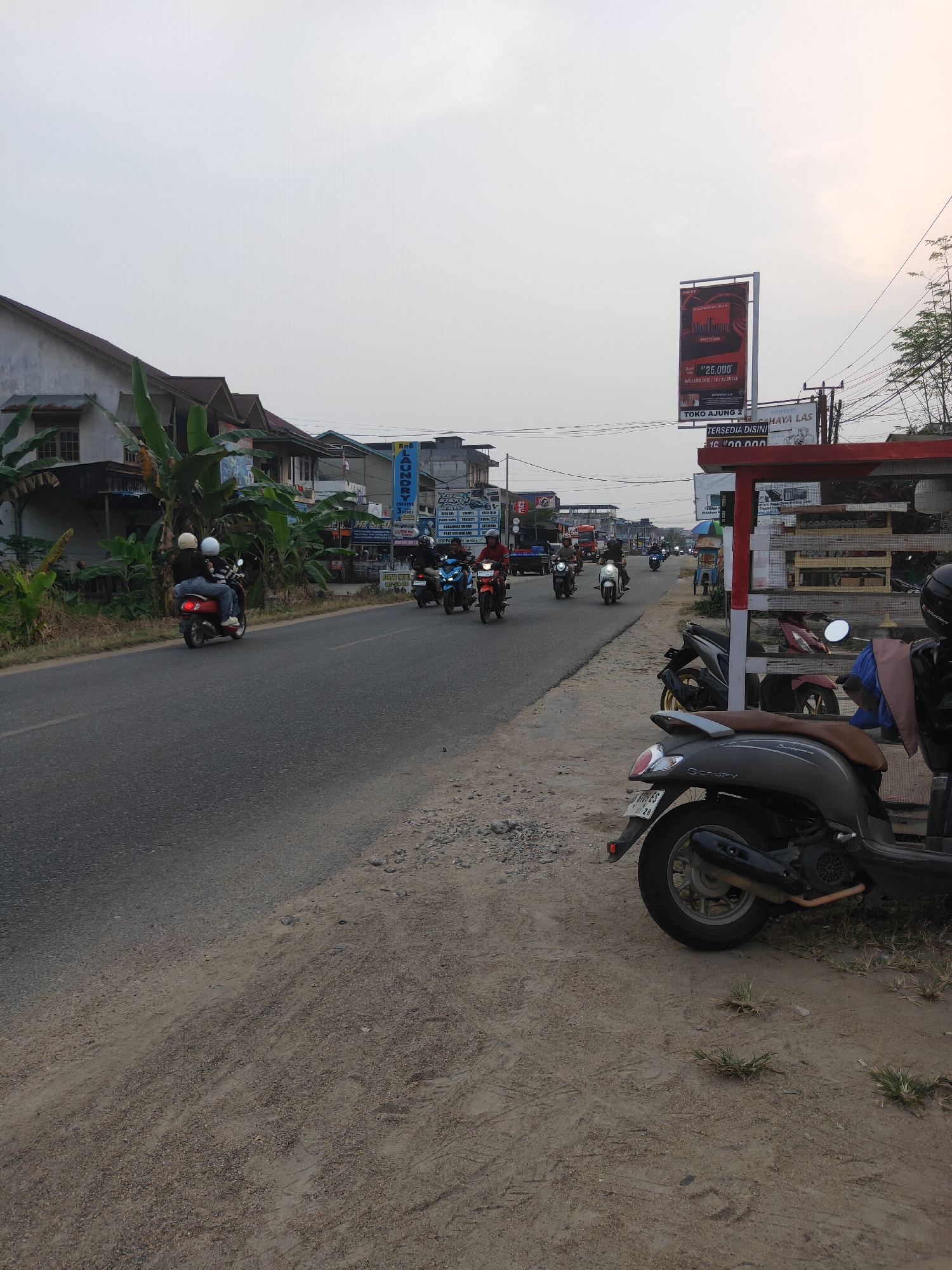 A street scene in Sintang, Indonesia, features a road with several motorcycles and scooters traveling in the same direction. The road is bordered by a dirt and gravel shoulder. On the left side of the road are buildings, including houses and shops, with some vegetation like banana trees. On the right side, there are more buildings, including a structure with shelves displaying items, and a billboard advertising "FILTER BLACK MOTION" cigarettes. The sky is overcast with the sun low on the horizon, casting a diffused light. A few people are visible riding the motorcycles, most wearing helmets. One scooter is parked in the foreground on the right.