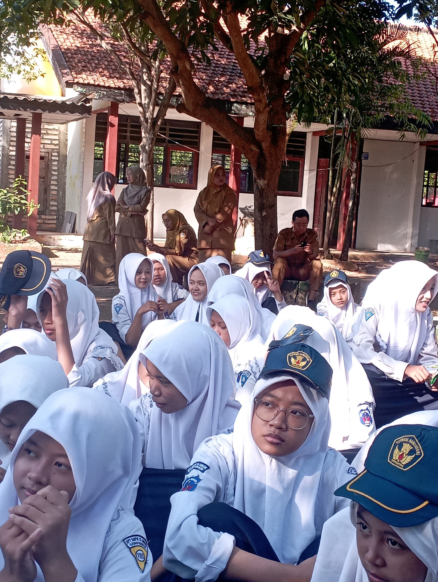 This image captures a group of students in uniform, likely at a school in Banyuurip, Indonesia. The students, predominantly female, are wearing white hijabs and uniforms, some with dark blue caps. They are seated outdoors, with a building visible in the background featuring a tiled roof and wooden window frames. Trees provide shade, with sunlight dappling through the leaves. In the background, two adults in uniform appear to be observing the students, one seated and looking at a phone. The overall scene suggests a casual gathering or activity taking place on school grounds.