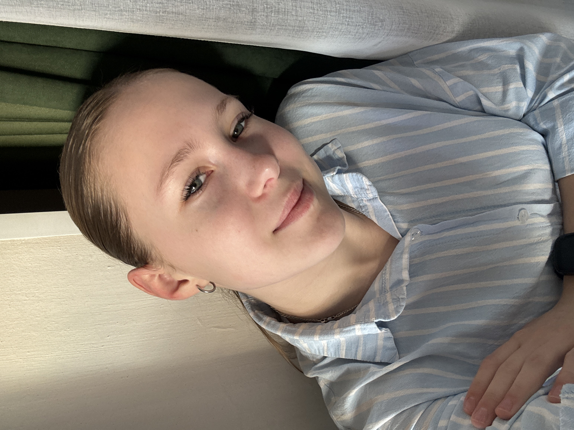 A young woman is featured in a close-up, angled portrait, captured indoors. She appears to be in her late teens or early twenties, with light-colored hair neatly pulled back from her face. Her light-colored eyes, possibly green or hazel, engage directly with the viewer, framed by visible eyelashes. Her expression is soft and pleasant, with a gentle smile gracing her lips, conveying a calm and content demeanor.
She is dressed in a light blue and white striped button-down shirt, possibly sleepwear or a casual top, with some buttons undone at the collar. A delicate silver chain necklace is visible around her neck, and a small silver hoop earring adorns her left ear. Her hands are resting on her body in the lower right of the frame, and a dark item, possibly a watch or fitness band, is visible on her right wrist.
The background is softly focused, suggesting an intimate indoor setting. Above her head is a textured, light-colored fabric, possibly a curtain or part of a bed, with a darker green fabric visible behind it. To her left, a light, subtly textured wall or surface is present. The lighting is soft and even, contributing to the tranquil atmosphere. The image captures a moment of relaxed comfort in what is understood to be Chaikovsky, Russia. There is no visible text in the image.