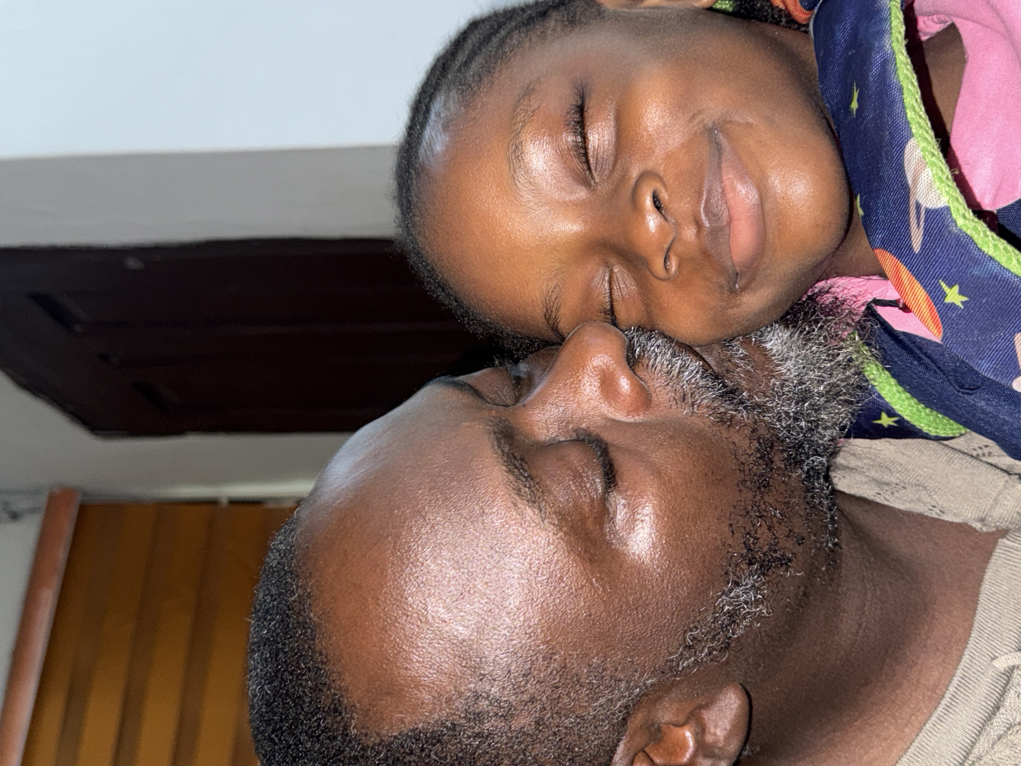 A close-up, rotated shot shows an adult male and a child sleeping. The child, possibly a girl, rests her head on the man's head and shoulder. Both have dark skin tones. The child wears a pink and blue patterned garment. The man has a short, graying beard and is wearing a tan shirt. The background reveals a white ceiling, a dark wooden panel or shelf, and the edge of a door frame. The location is possibly Ado, Nigeria, based on provided context.