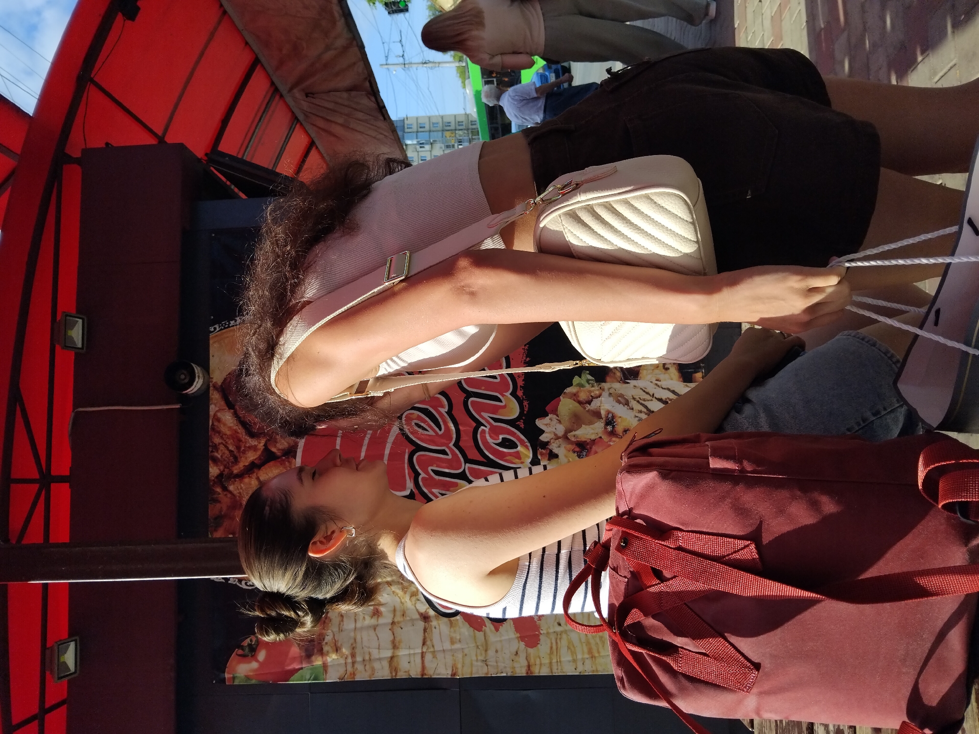 Two young women are standing outdoors next to a food stall with a red awning and a large banner depicting food items. The woman on the left, with her hair in a bun, is wearing a striped tank top and denim shorts. She has a burgundy backpack on the ground in front of her. The woman on the right is wearing a light-colored crop top and dark shorts and has a cream-colored quilted crossbody bag. She is leaning forward, facing the other woman, and her face is obscured. In the background, there is a street scene with a green bus, a blue car, and two other individuals walking. The environment appears to be a public area in the city of Zhytomyr, Ukraine.