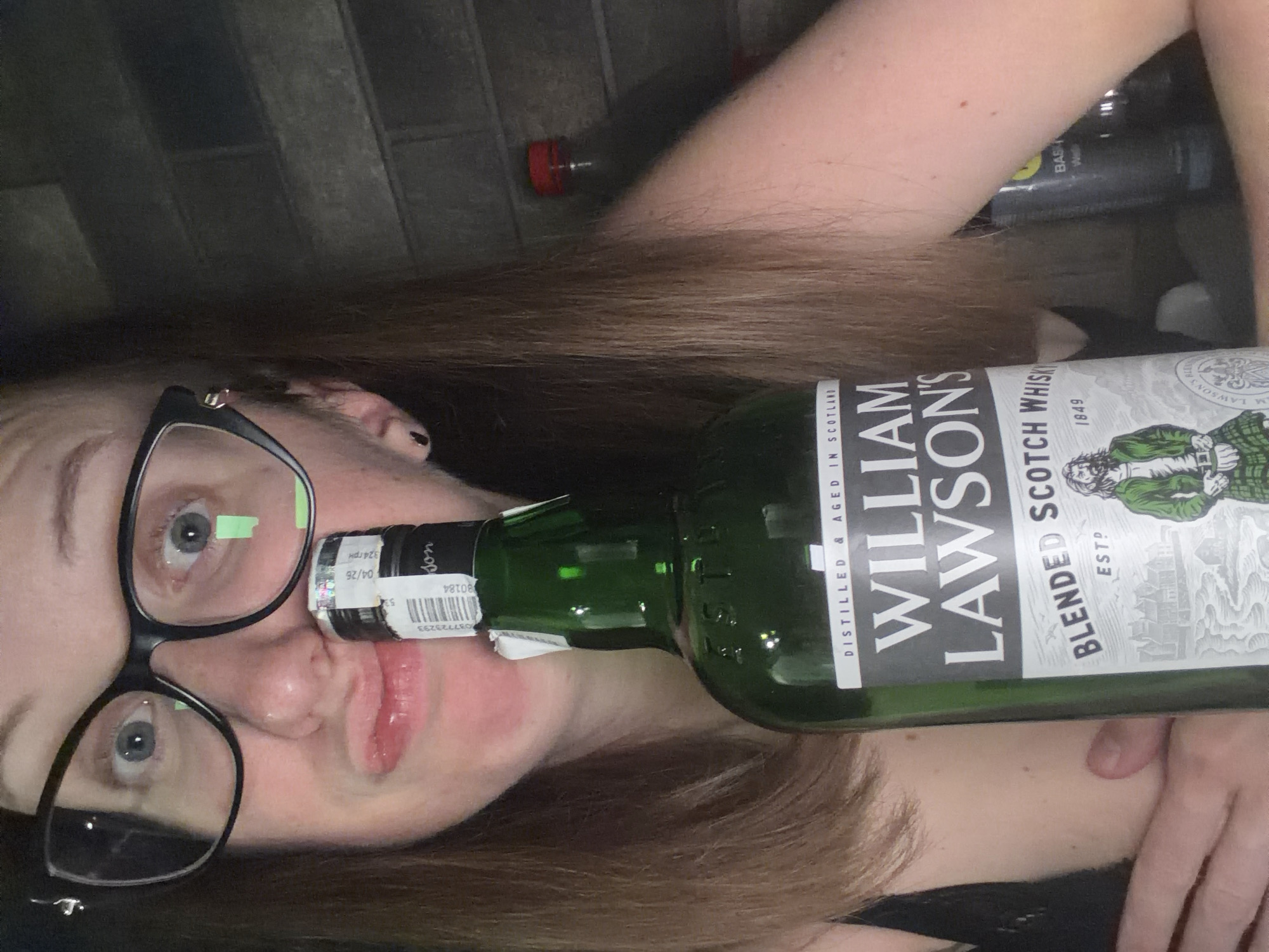 A person with light brown hair and glasses is holding a bottle of William Lawson's blended Scotch whisky. The person's face is close to the camera, with their lips pursed. The background appears to be a brick wall. There is a red-capped bottle in the background. There is no indication of location in Bila Tserkva, Ukraine.