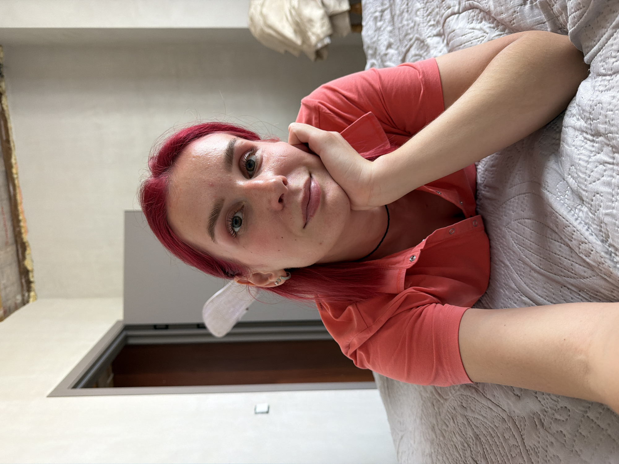 An image captures a woman posing for a selfie while lying on a bed in a room located in Chaikovsky, Russia. The woman has fair skin, reddish-pink hair, and is wearing a coral-colored short-sleeved collared shirt with snap buttons. She wears a thin black choker necklace and multiple earrings on her left ear, including a hoop, a stud, and a small gem stud. Her left hand is positioned under her chin, propping her head up, while her right arm extends out of frame to the lower left. She is looking directly at the camera with a slight smile. The bed features a light grey or beige quilted bedspread with a repetitive geometric pattern. In the background, white walls are visible. To the left, a doorway with a grey frame reveals a dark brown door beyond it. Above the doorway and extending into the ceiling, exposed ceiling joists and insulation material are visible. A small, indistinct white object, possibly a hair accessory, is positioned behind her head to the left. To the right of the woman, a piece of furniture draped with light-colored fabric is partially obscured. Natural light illuminates the scene, suggesting an indoor setting during daylight hours.
