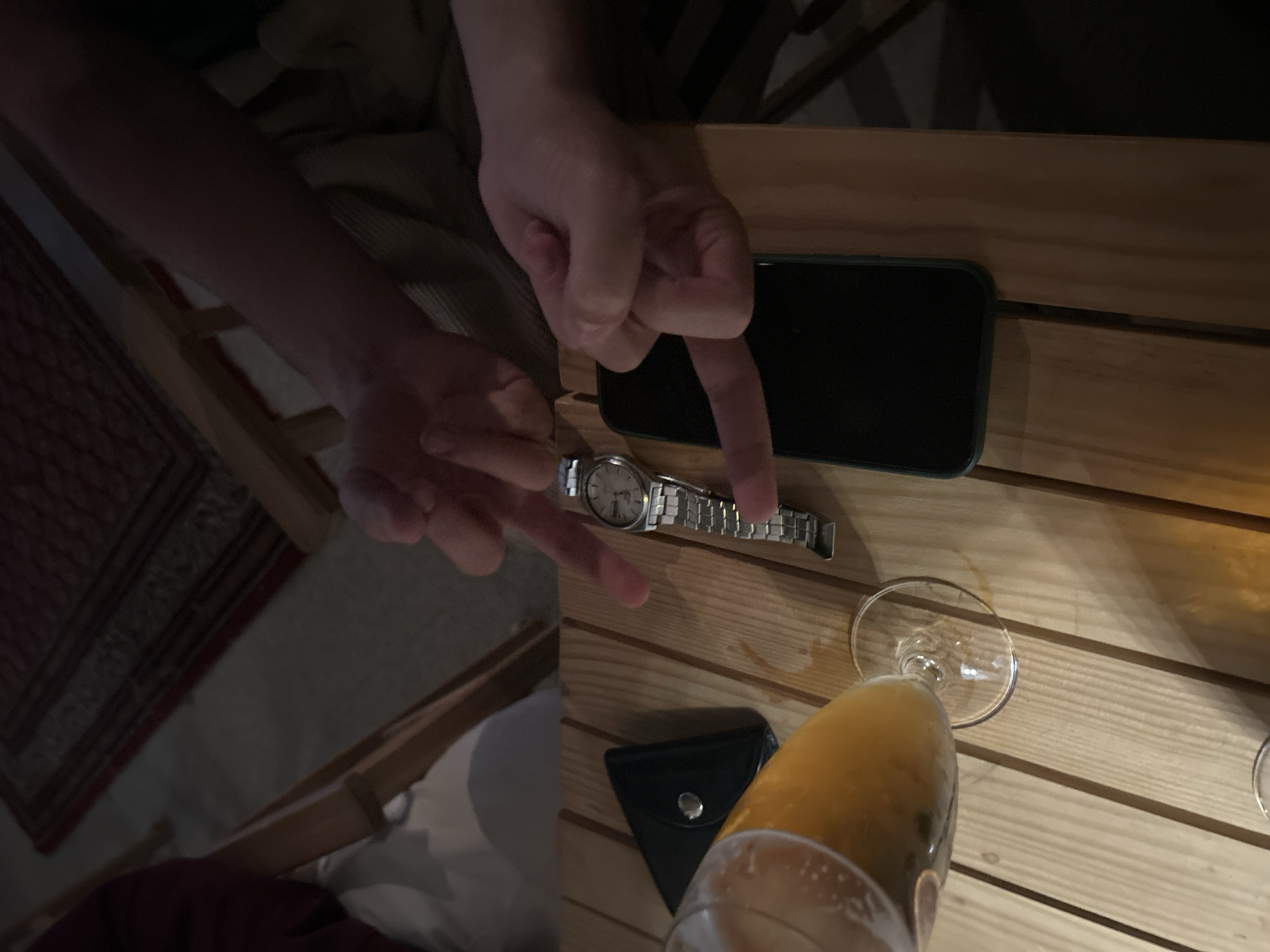 A view of a wooden slat table holding a black smartphone, a silver wristwatch with a round face, a small black leather wallet, and a glass of beer. Two hands are reaching toward the watch, apparently interacting with it. The scene is indoors, likely at a bar or similar establishment in Mirandela, Portugal. The light conditions indicate it may be nighttime. Parts of a chair and a rug are also visible in the background.