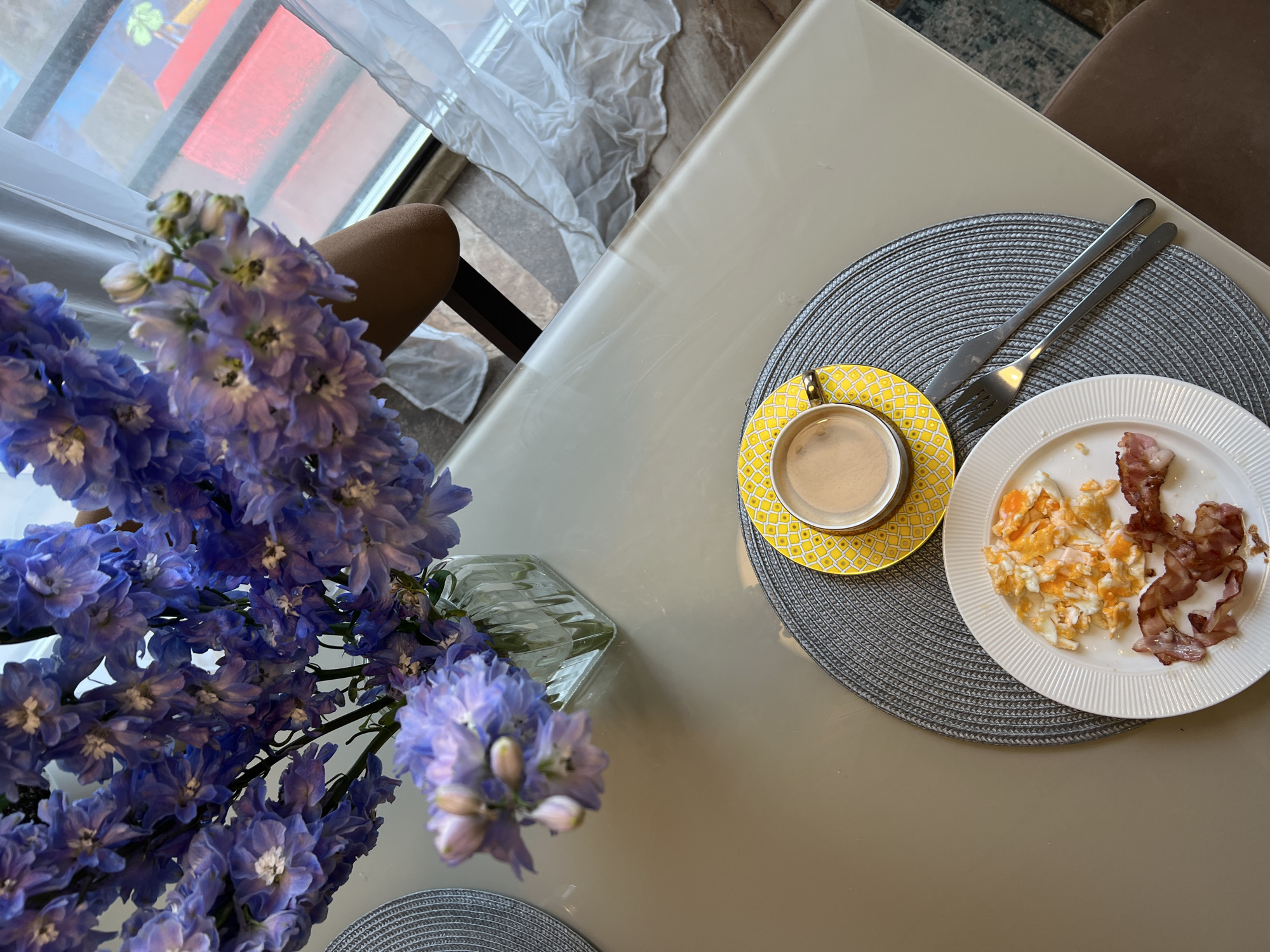 The image displays an overhead view of a breakfast setting on a light-colored, possibly glass, table in Khimki, Russia. To the left, a lush bouquet of purple flowers, likely Delphiniums, stands in a clear glass vase, adding a vibrant natural element to the scene.
On the right side of the table, a single place setting is arranged on a circular gray woven placemat. A bright yellow teacup with a distinctive geometric pattern rests on a matching saucer, containing a beverage, possibly coffee or espresso. Next to the cup is a white plate, designed with subtle vertical ridges, holding a portion of scrambled eggs and several strips of bacon. A silver fork and knife are neatly placed on the placemat beside the plate.
In the upper left background, a large window is visible, letting in abundant natural light. A sheer white curtain hangs near the window, and indistinct reflections, including hints of red, are visible through the glass, suggesting an outdoor view. Part of a brown chair back can also be seen near the window. The scene depicts a quiet and well-lit morning meal arrangement. No people are present, and there is no visible text in the image.