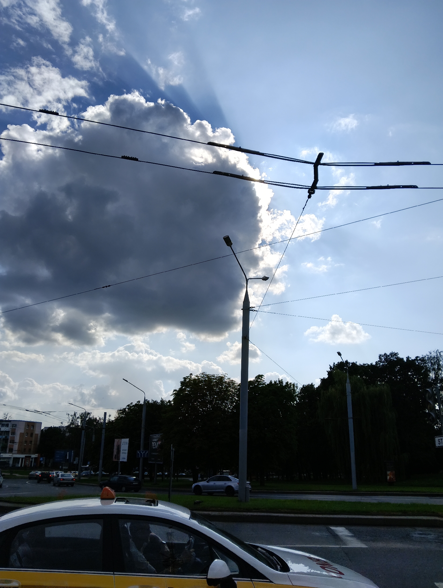 The image depicts a street scene in Hrodna, Belarus, on a partly cloudy day. The sky is dominated by a large, dark cloud with sunbeams emanating from behind it. Overhead, trolleybus wires crisscross the frame, supported by poles. In the foreground, the roof and hood of a white taxi with a yellow stripe are visible. The taxi has an orange light on its roof. The road has lane markings and a pedestrian crossing. Beyond the road, a grassy median separates the traffic from a sidewalk lined with trees. In the background, there are buildings and more streetlights. The overall impression is of a typical urban environment with a dramatic sky.