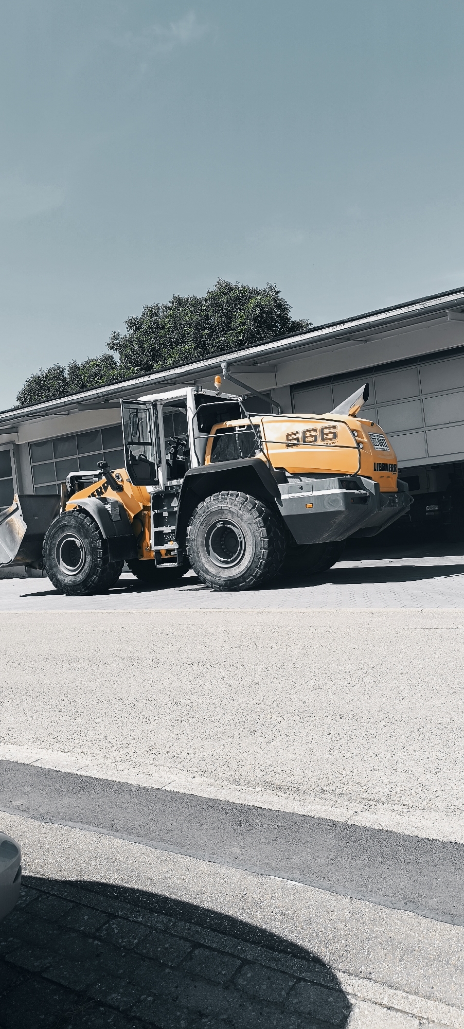 A yellow and gray Liebherr wheel loader with the number "586" on its side is parked on a paved surface in front of a building with a large garage door. The building appears to be a commercial or industrial structure with a white facade and a dark roof. Trees with green foliage are visible behind the building, and a clear blue sky with a few wispy clouds is overhead. The wheel loader has large, rugged tires and a scoop at the front. The scene is well-lit, suggesting it is daytime. The image appears to have been taken outdoors, likely in a yard or lot associated with the building. The overall impression is of a quiet, static scene, with the heavy machinery at rest. It is difficult to determine the exact location within Winterbach, Germany, but it is likely in an industrial or commercial zone. There are no people or animals visible in the image, nor any signs of recent activity.