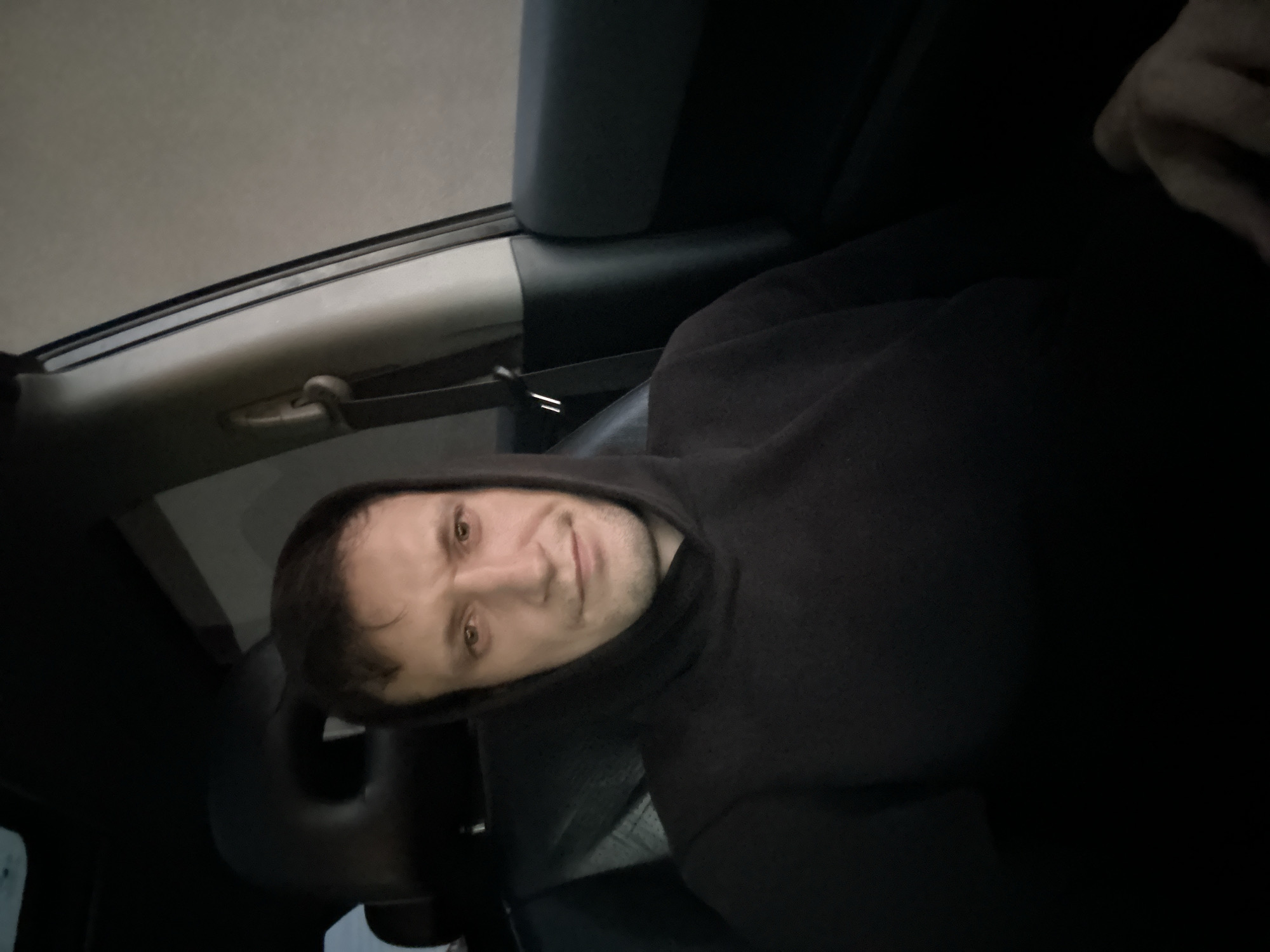 A medium shot of a man with short brown hair wearing a black hooded sweatshirt, seated in the passenger seat of a car. The man is looking directly at the camera, with a neutral expression. The background shows the interior of the car, including a headrest and part of the door panel. The lighting is dim, suggesting it is either nighttime or the interior of the car is poorly lit. The image does not provide any information about the location of Shatava, Ukraine.