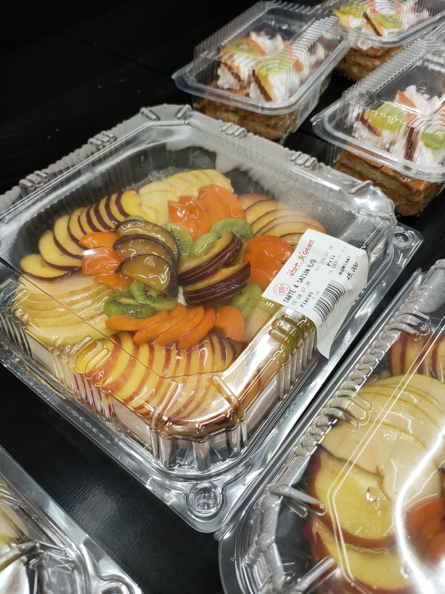 A close-up, high-angle shot shows a clear plastic container filled with sliced fruit, primarily peaches and possibly apples, along with slices of kiwi and apricot. The fruit is arranged in a circular pattern on top of a pastry base, covered in a clear glaze. A label on the side of the container reads "Tarte 4 Saison G/B" and "DULC.09 07 26", indicating it is a seasonal tart with a date of July 26th. Other nearby containers also hold pastries, some topped with whipped cream and fruit. The containers are placed on a dark, reflective surface. The lighting suggests an indoor environment, possibly a bakery or supermarket display. The image was taken in Cebalat, Tunisia, as indicated by the context.
