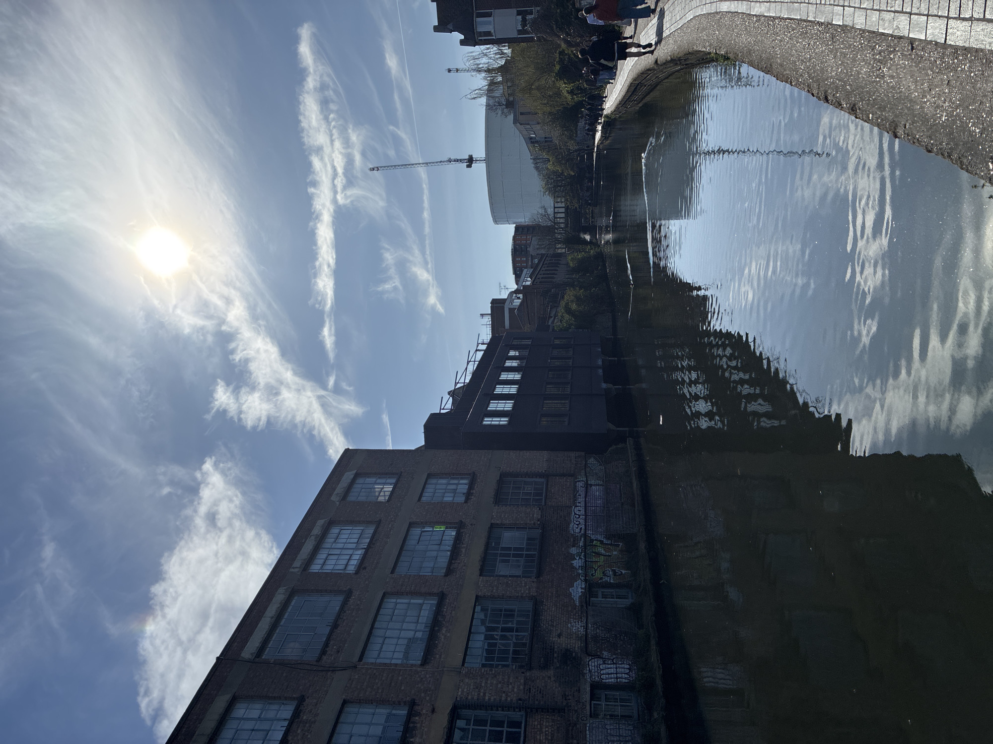 The photograph depicts a canal in London, United Kingdom. On the bank of the canal, figures are seen walking. A brick building rises on one side, reflecting in the water. Graffiti marks a section of the building's wall. A construction crane is visible in the distance, adjacent to a large, cylindrical structure. The sky is a bright blue with thin, wispy clouds, and the sun shines brightly, creating glare in the image.