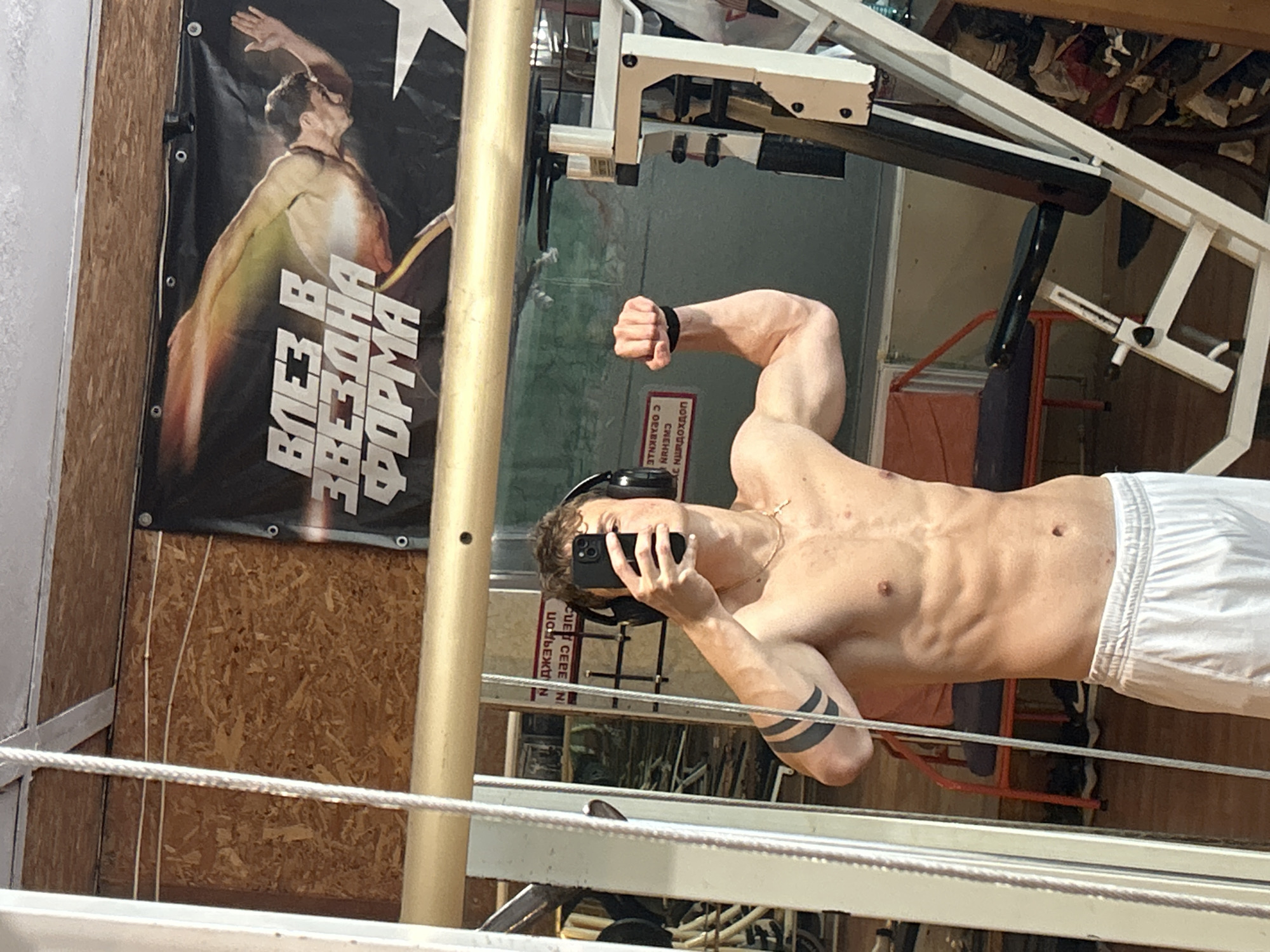 A partially visible shirtless man with white shorts and two black bands tattooed on his right arm is taking a selfie with his phone in a mirror in Elhovo, Bulgaria. He is wearing headphones. Behind him, on a wooden wall, there is a poster with a shirtless man and text written in Cyrillic. The surrounding environment includes a metal structure that could be part of a gym or fitness area.
